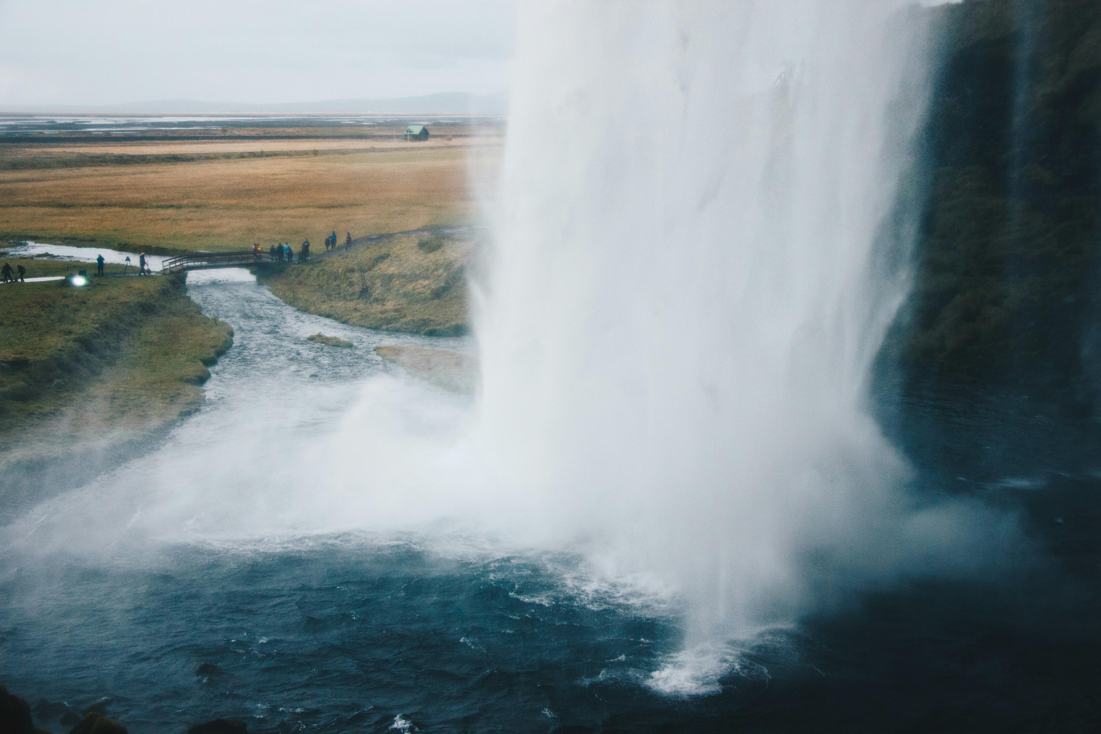 Seljalandsfoss Waterfall in Iceland, capturing the powerful water stream flowing into a river, surrounded by fields and distant visitors.