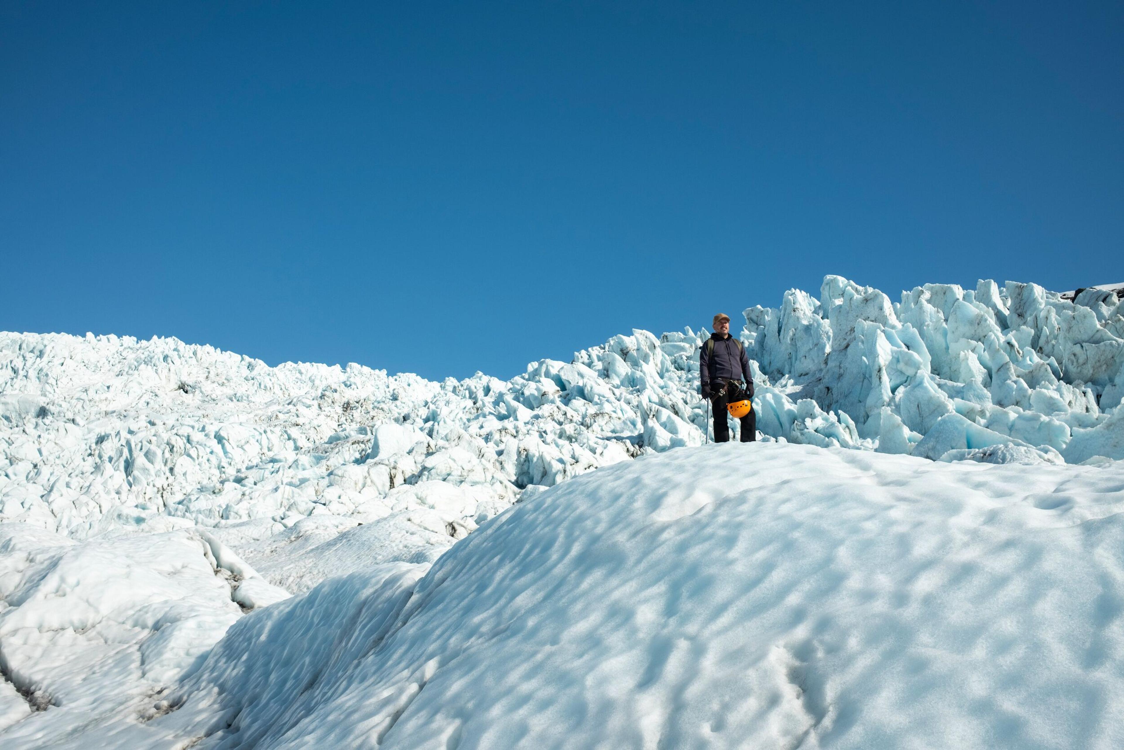 A person holding an orange helmet stands on a vast glacier under a clear blue sky.