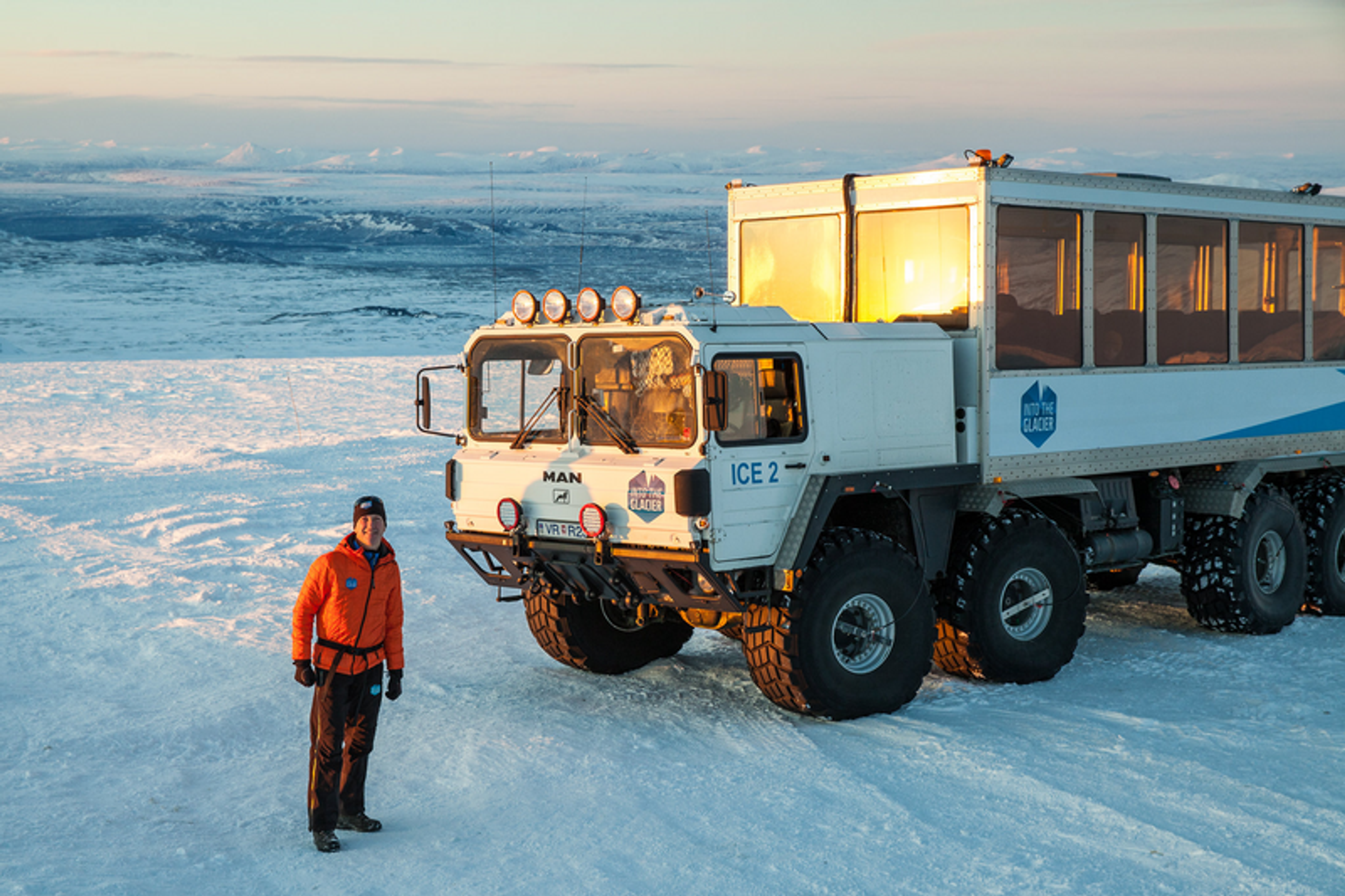 A person in an orange jacket stands beside a large, rugged glacier truck labeled "ICE 2" on the snowy terrain of Langjökull, with a scenic view of Iceland's icy landscape at sunset.