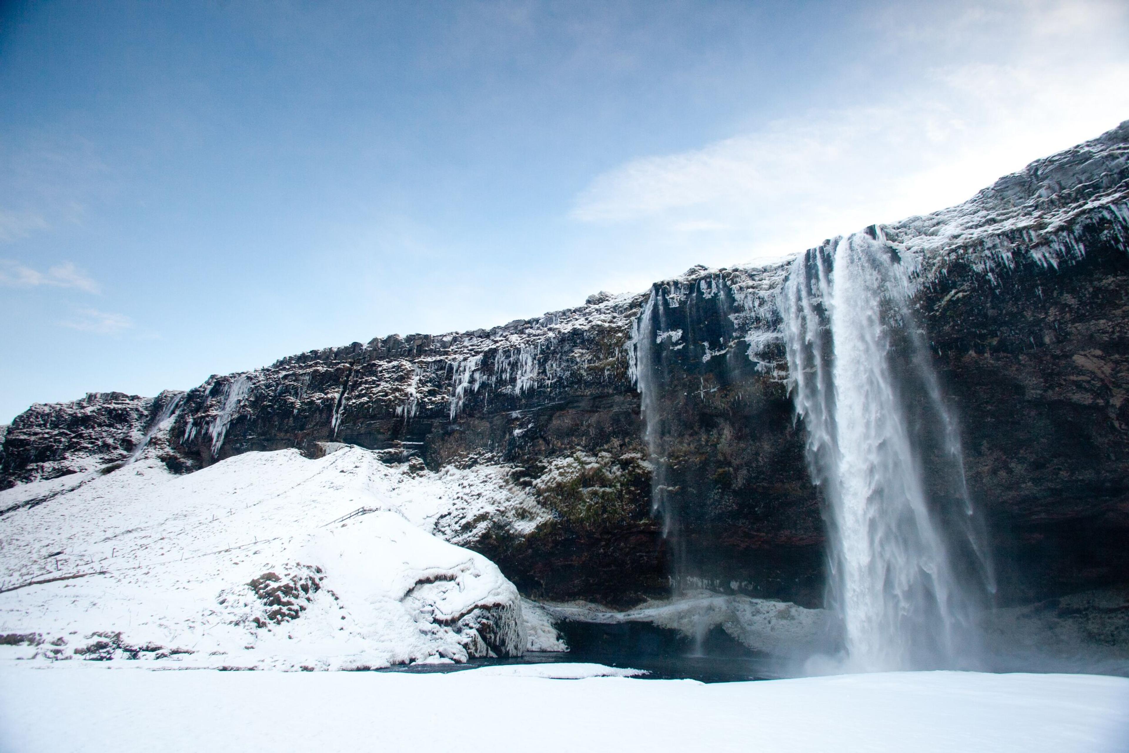 A snowy scene with a powerful waterfall plunging over a cliff against a clear blue sky, with ice and snow covering the surrounding landscape.