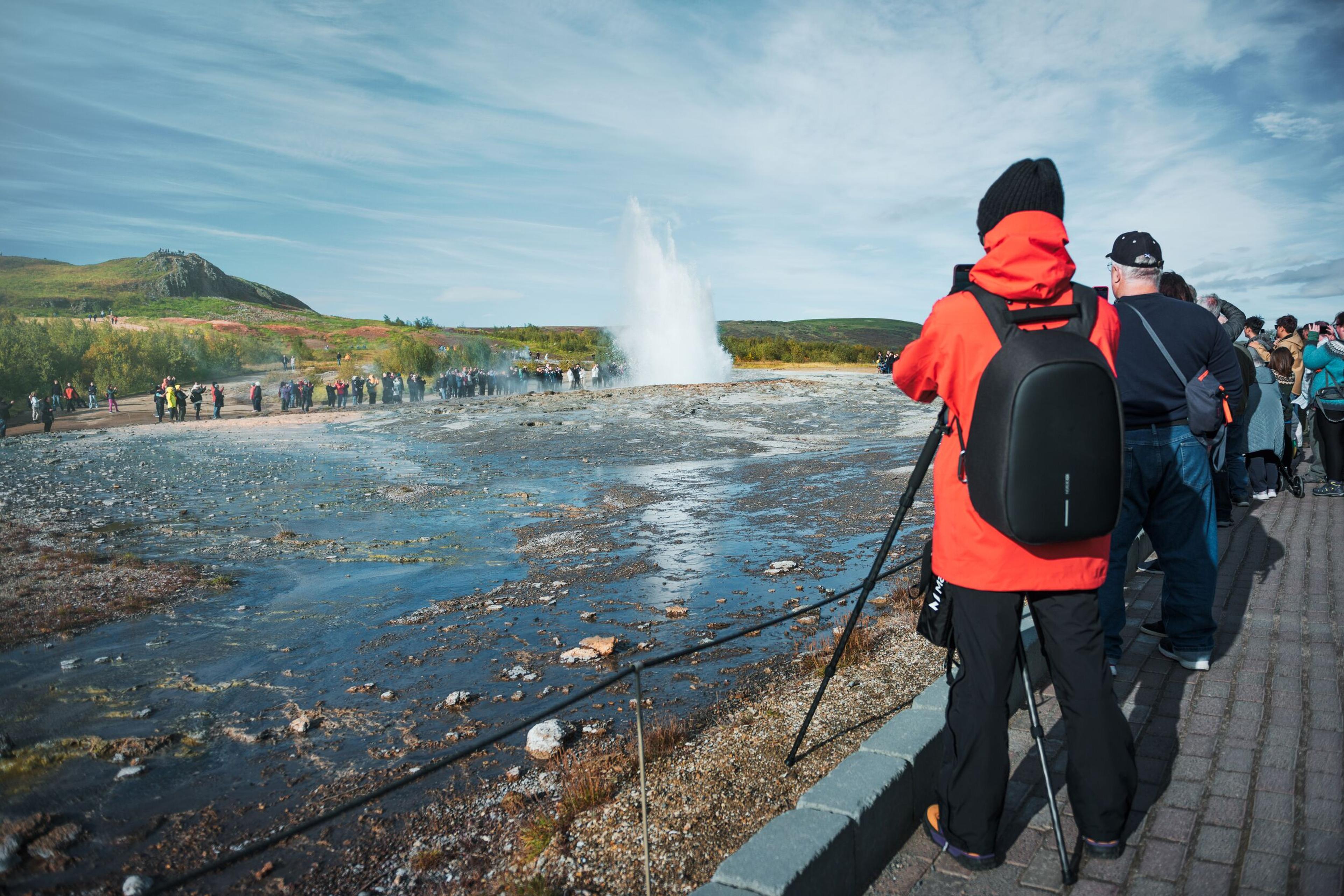 A visitor clad in a red jacket with a camera on a tripod observes a geyser erupting in Iceland, while a crowd of tourists at a distance watches the natural spectacle under a clear blue sky.