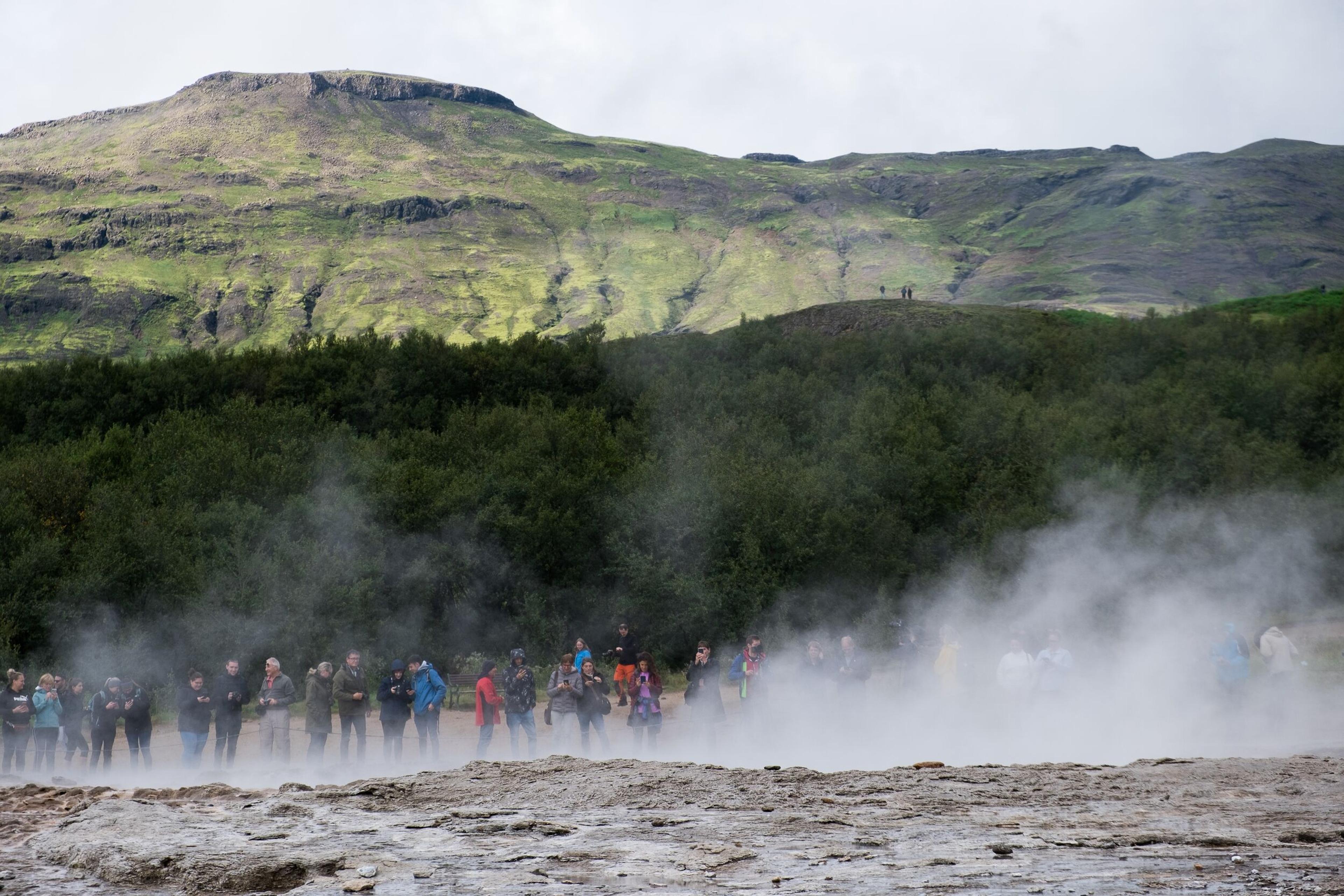 Tourists standing in steam at the Geysir geothermal area in Iceland, with a lush green hill in the background.