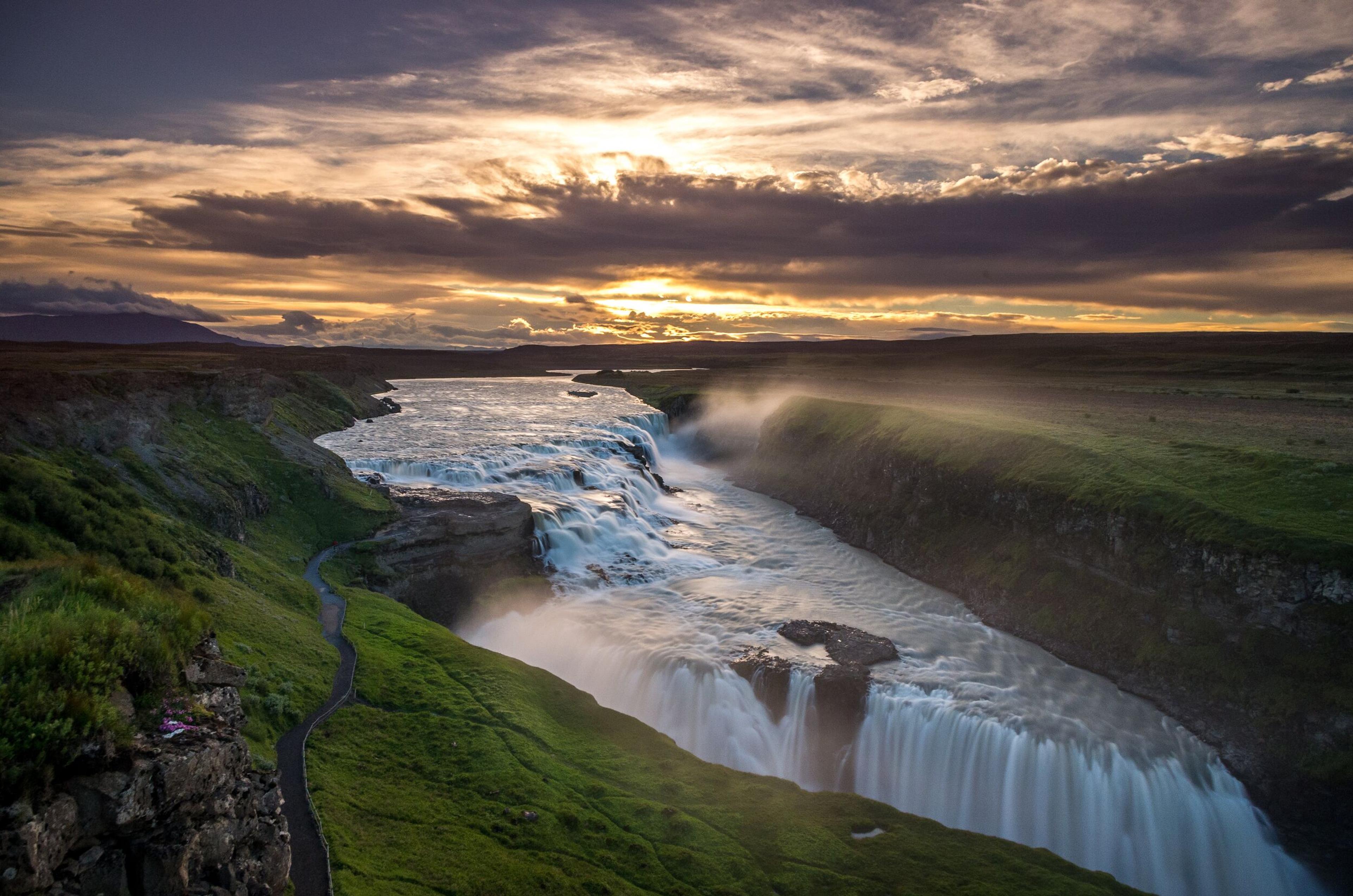 A stunning waterfall cascades through a lush green valley under a dramatic cloudy sunset. The golden light enhances the contrast between the water and the surrounding landscape.