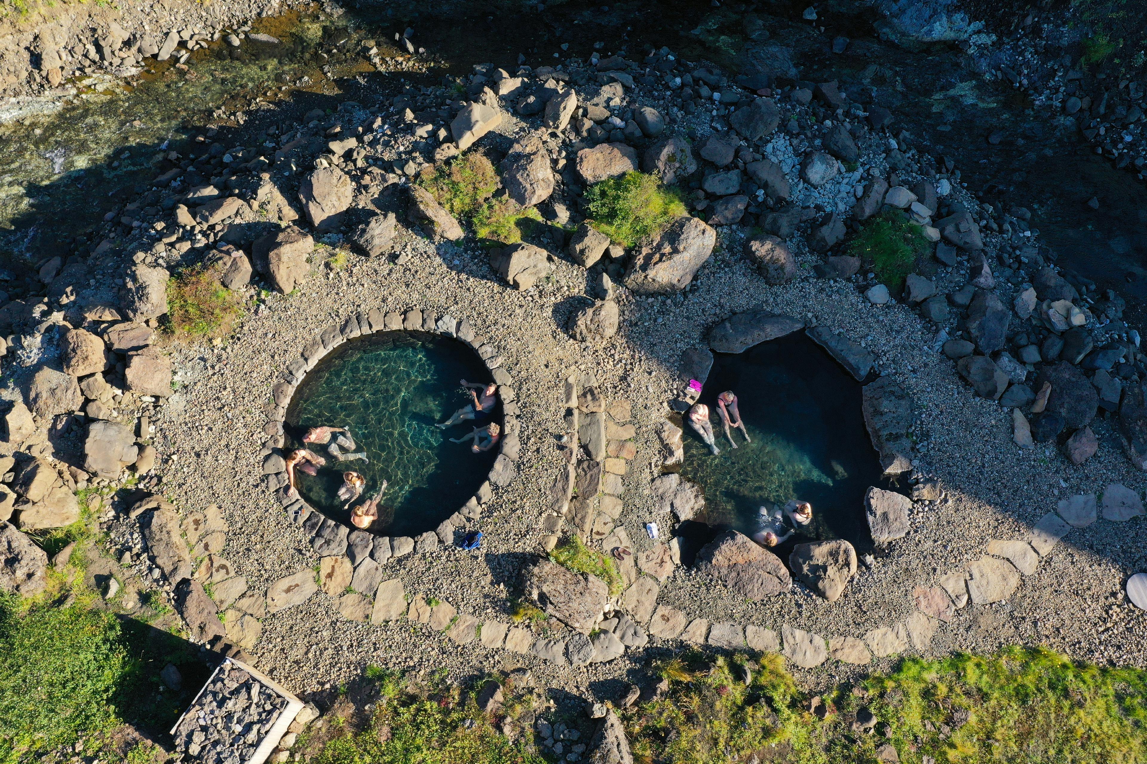 Aerial view of natural hot springs in Húsafell, featuring two circular stone pools surrounded by rocks and greenery, with people relaxing in the geothermal waters.