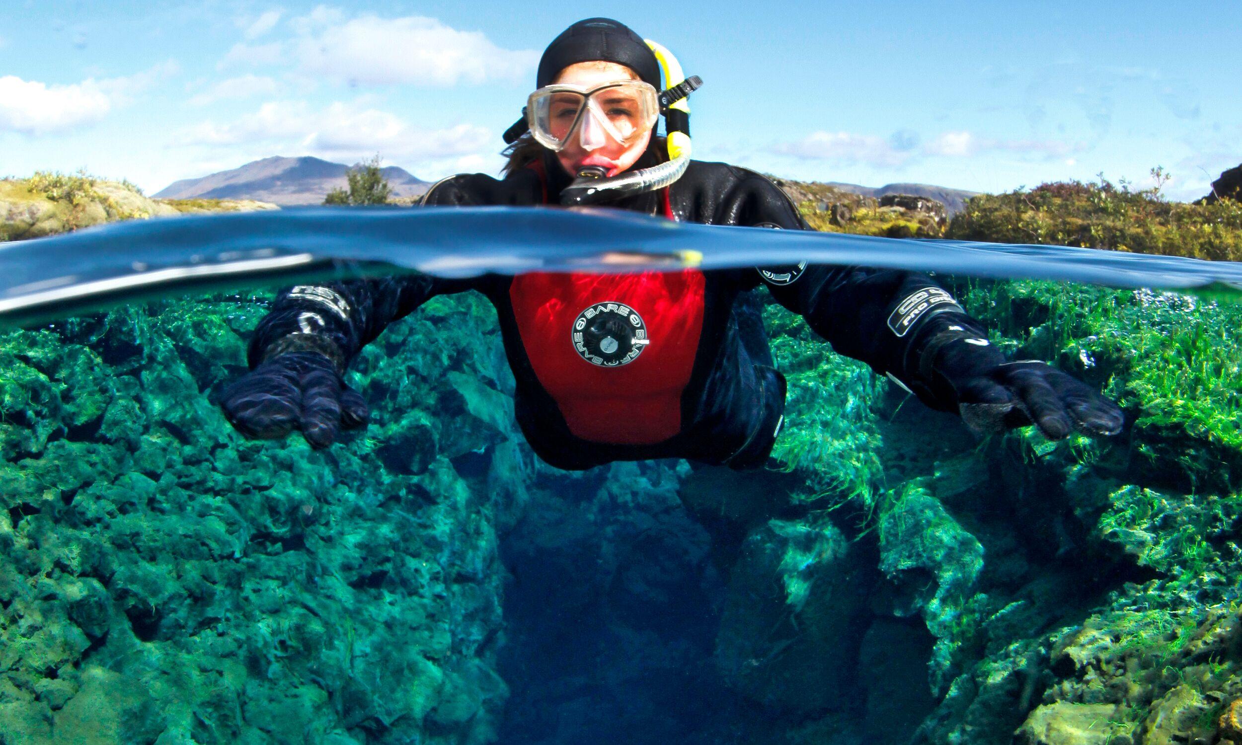 Snorkeler in a dry suit floating in the crystal-clear waters of Silfra fissure, surrounded by vibrant underwater rock formations.
