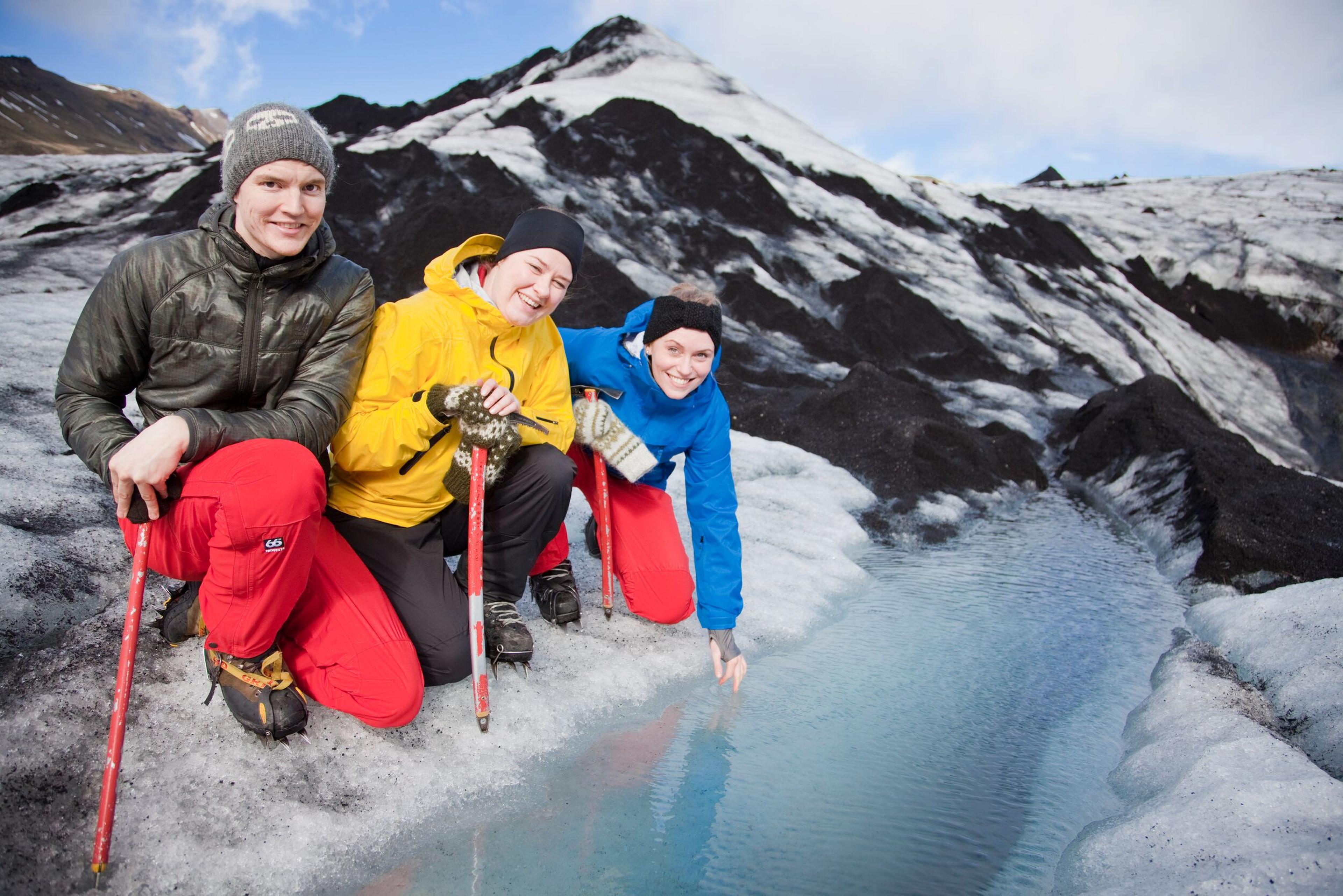 hree hikers in colorful jackets smile as they touch the crystal-clear meltwater on Sólheimajökull glacier, surrounded by volcanic ash and ice. A refreshing moment during a Sólheimajökull glacier hike in Iceland.