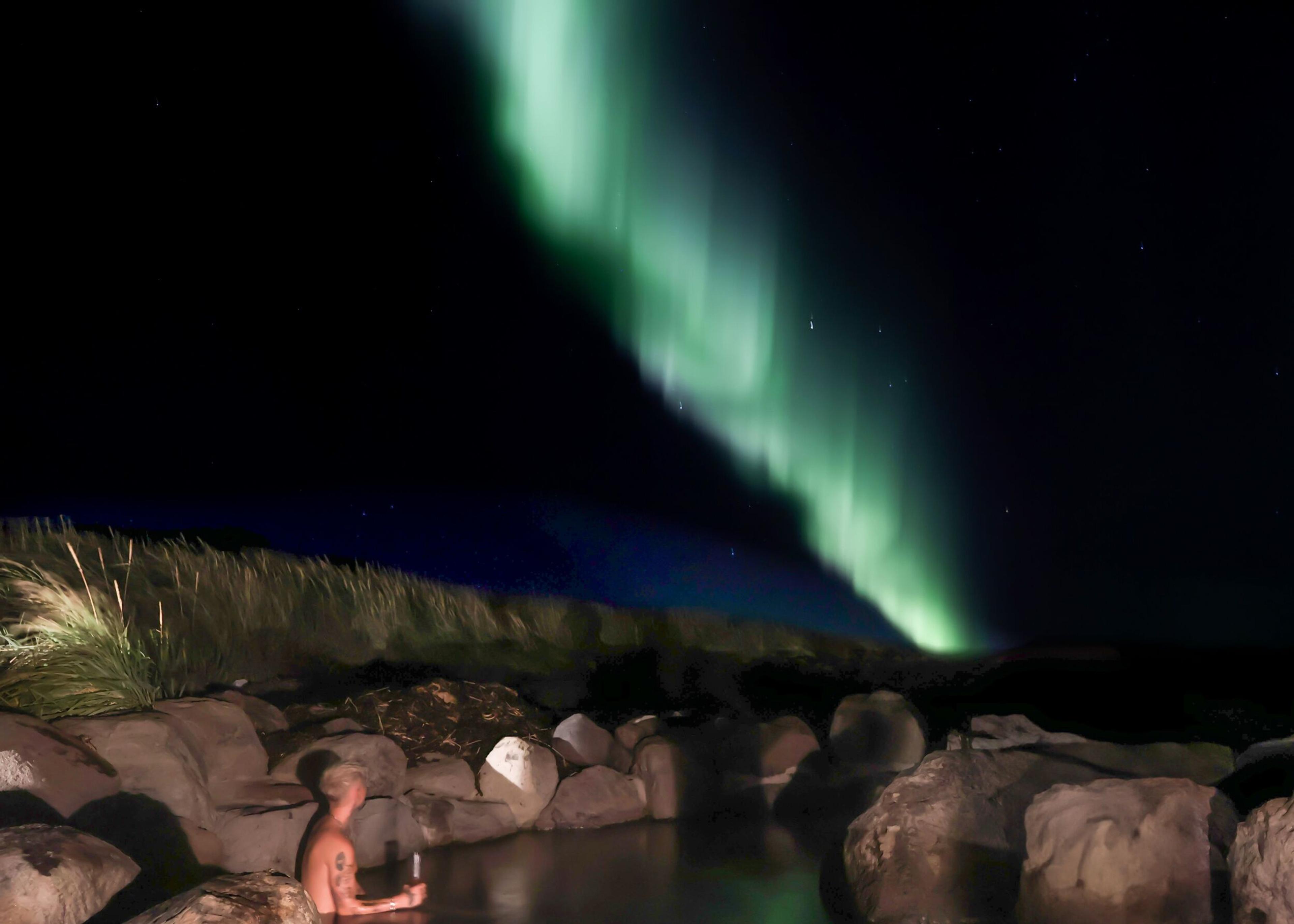 A man unwinds in a steaming hot spring, gazing upwards at the vibrant Northern Lights shimmering across the night sky while sipping a drink
