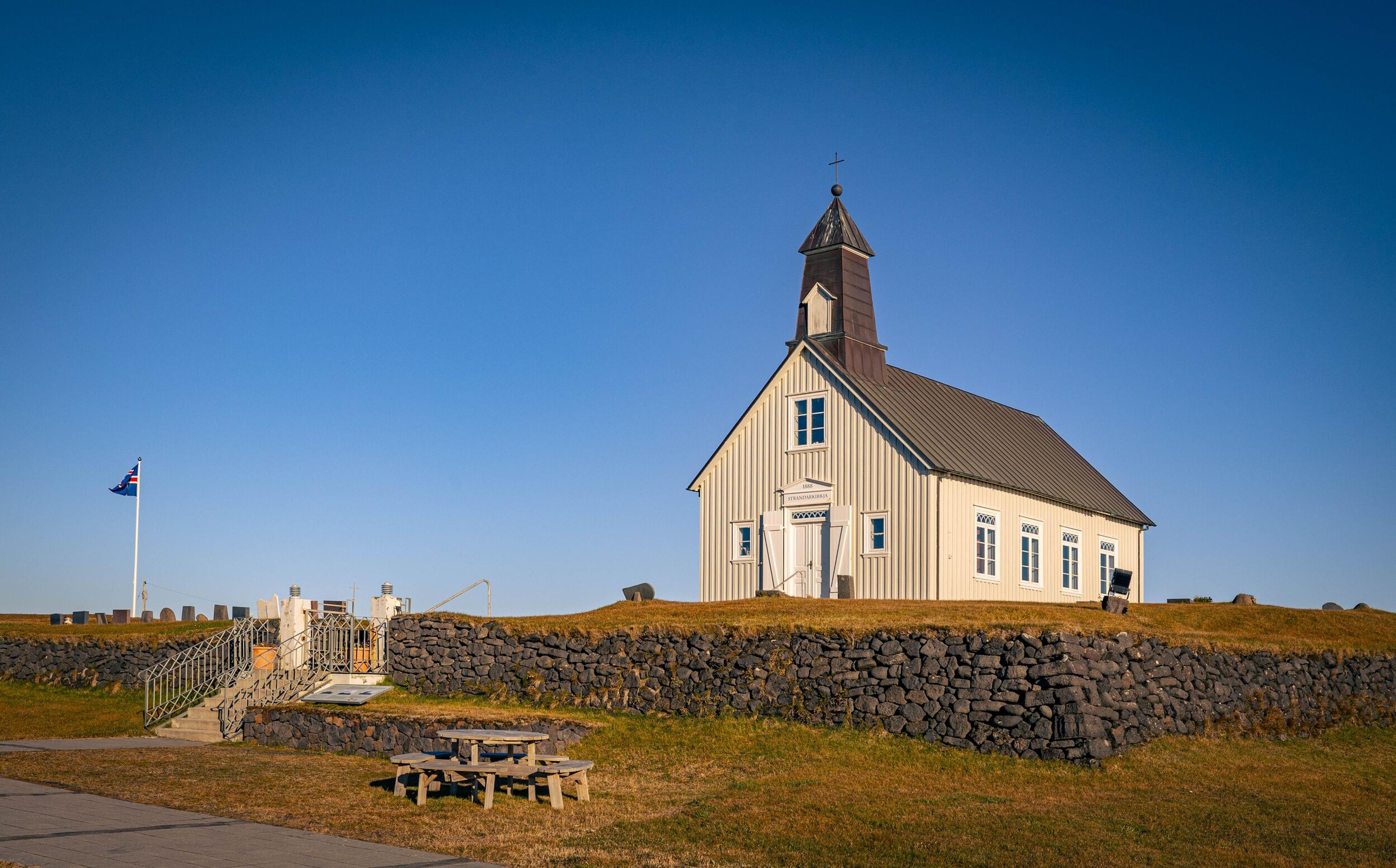 Traditional wooden church with a steeple in a countryside setting, bordered by stone walls and clear blue skies above.