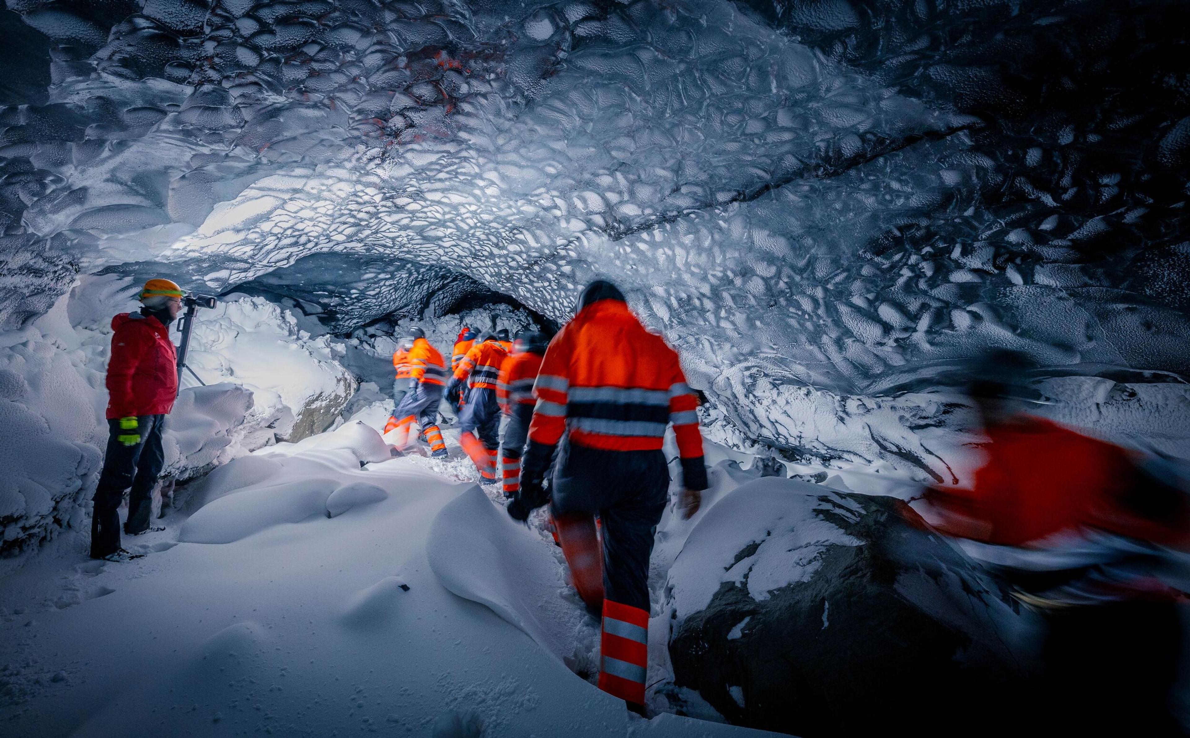 The image captures a group of explorers wearing high-visibility orange and red jackets with reflective stripes, navigating through a snow-filled ice cave