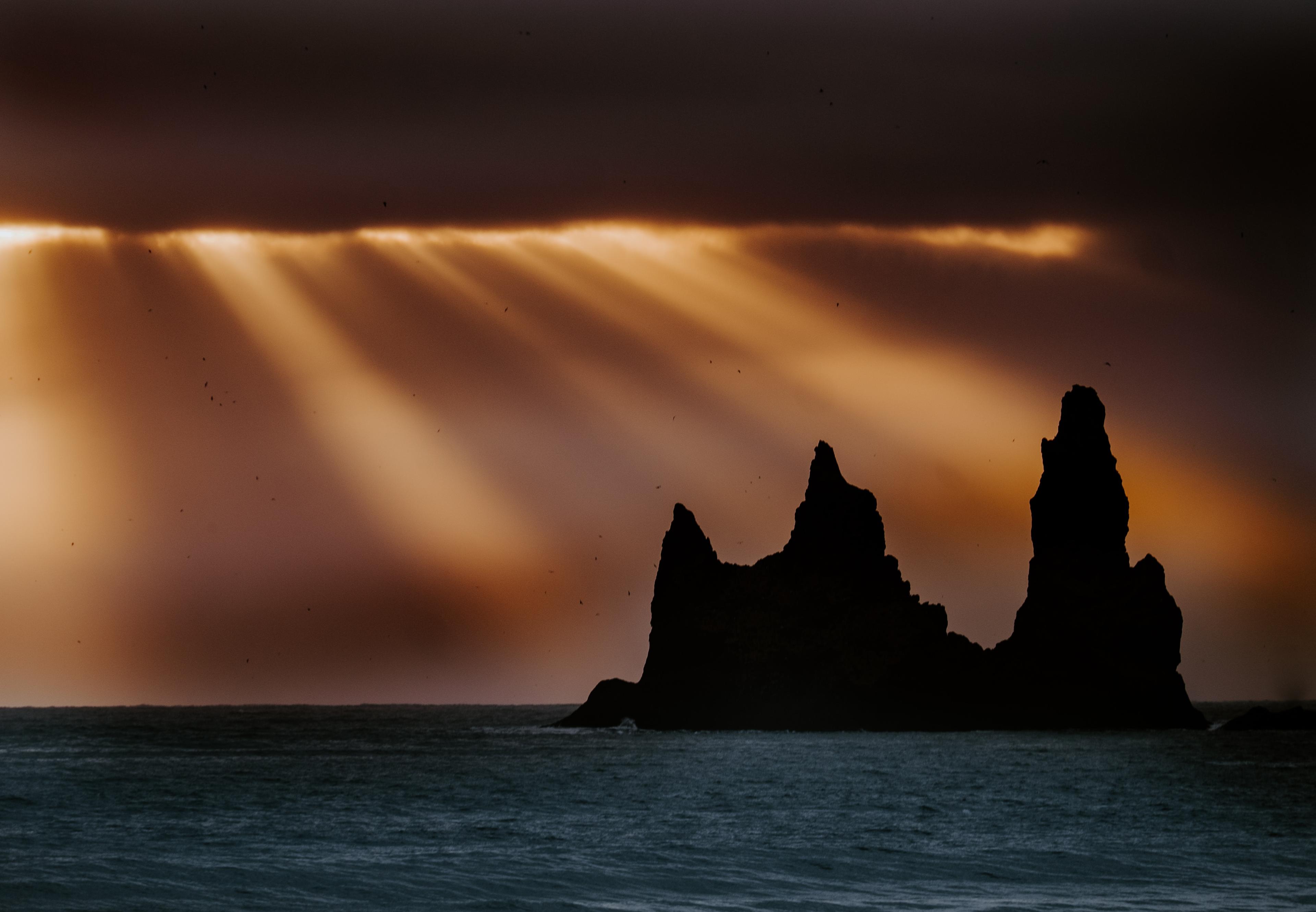 The silhouette of Reynisdrangar sea stacks set against a dramatic sky, where radiant sunset rays pierce through dense clouds, casting fiery hues across the horizon.