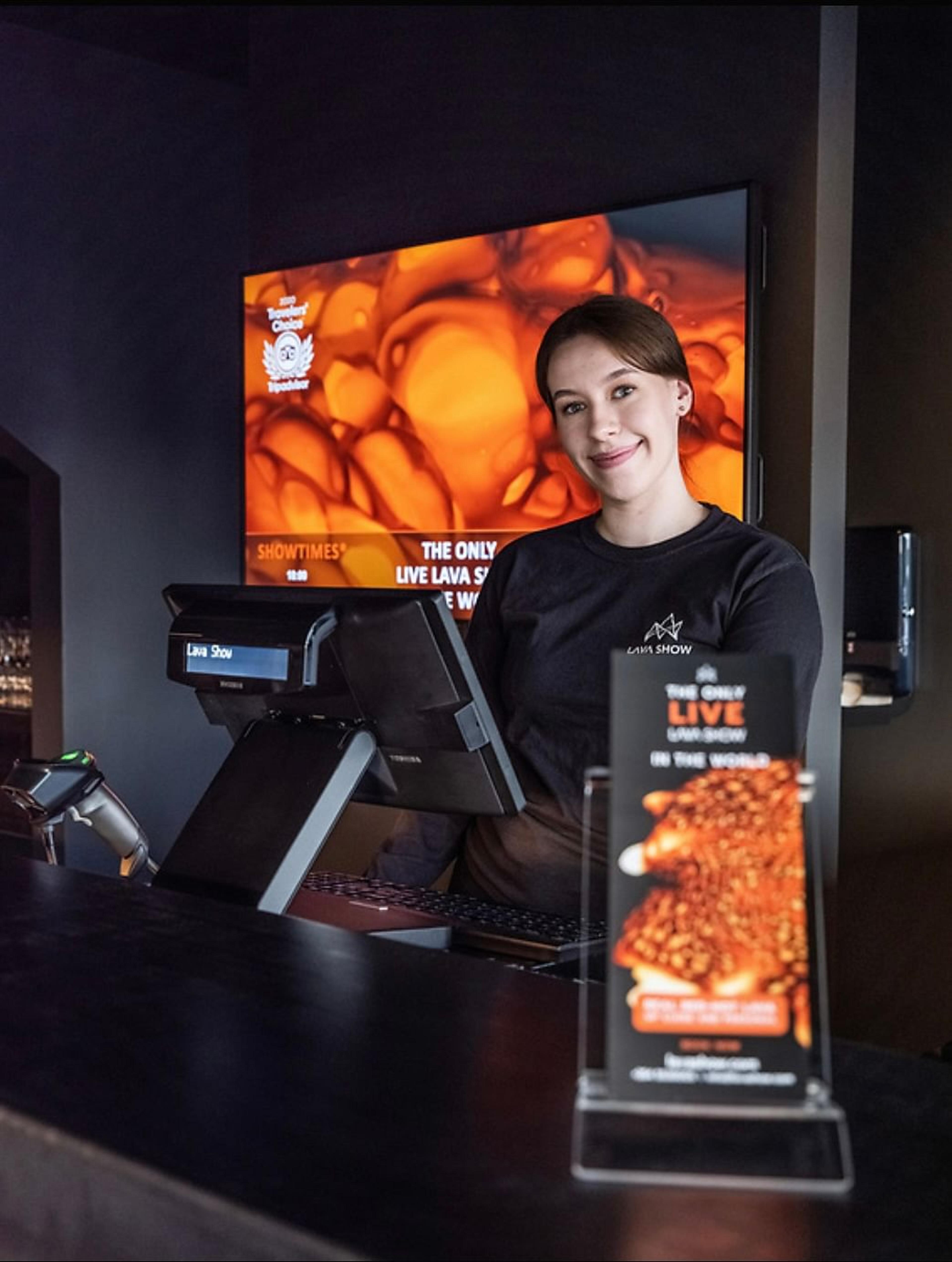 A smiling woman in a black "Lava Show" shirt stands behind a counter with POS systems, in front of a large screen displaying orange lava and "Showtimes" for "The Only Live Lava Show."