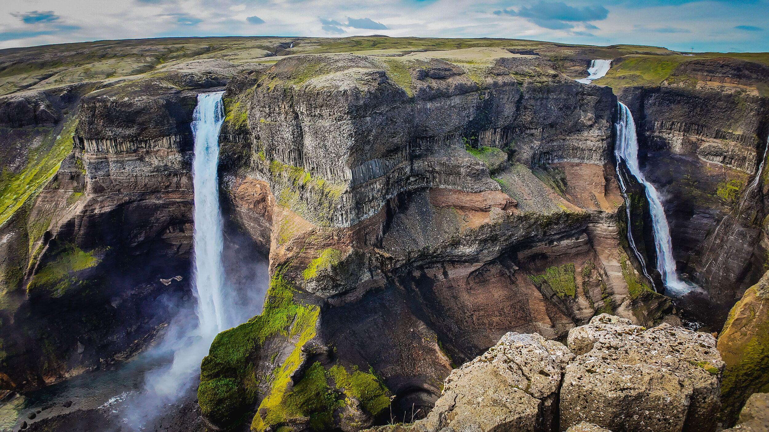 Two waterfalls cascade into a deep, rocky canyon with green moss under a partly cloudy sky.