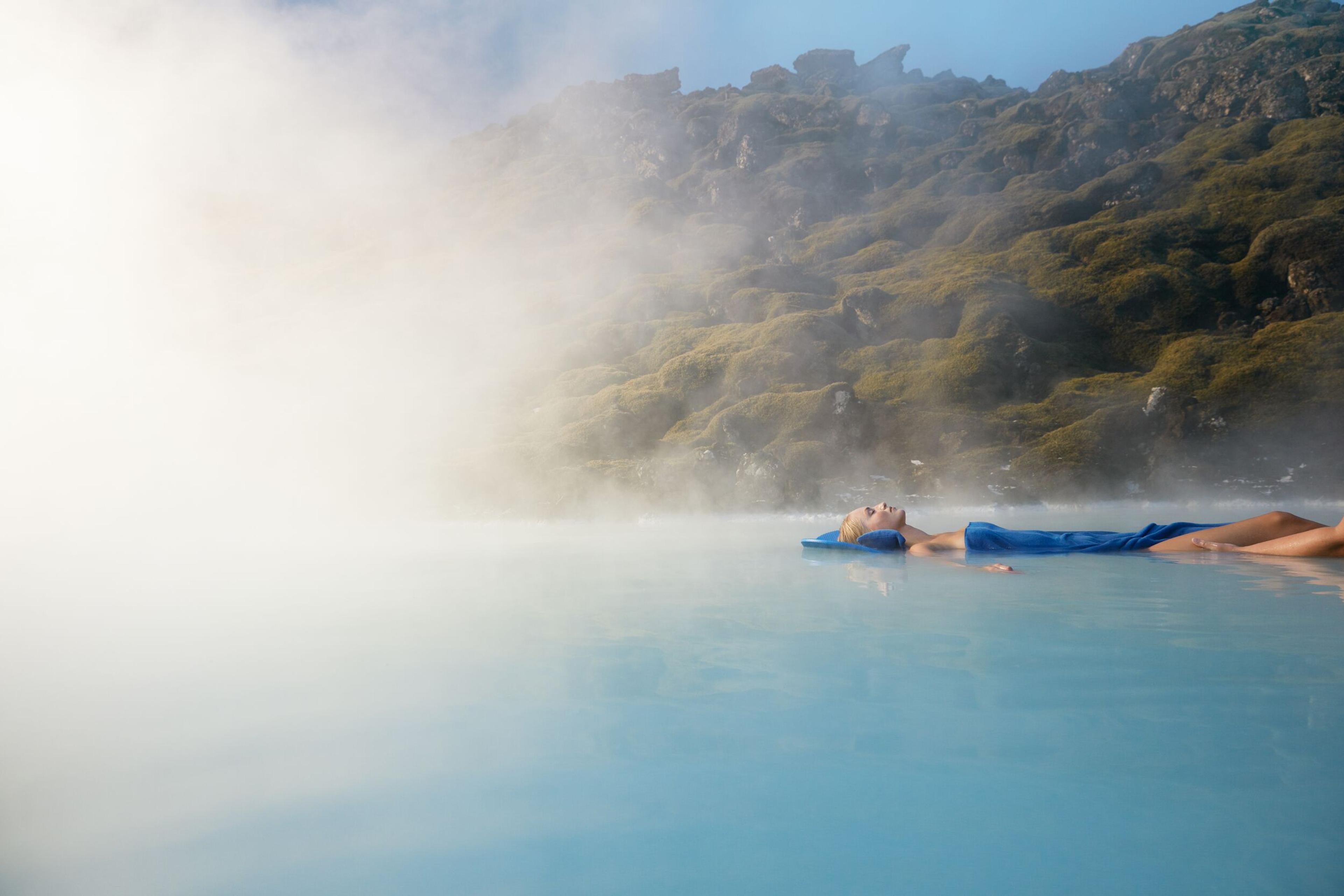 A woman draped in a blue towel floats serenely in the Blue Lagoon, enveloped by rising steam, with verdant moss-covered rocks in the backdrop
