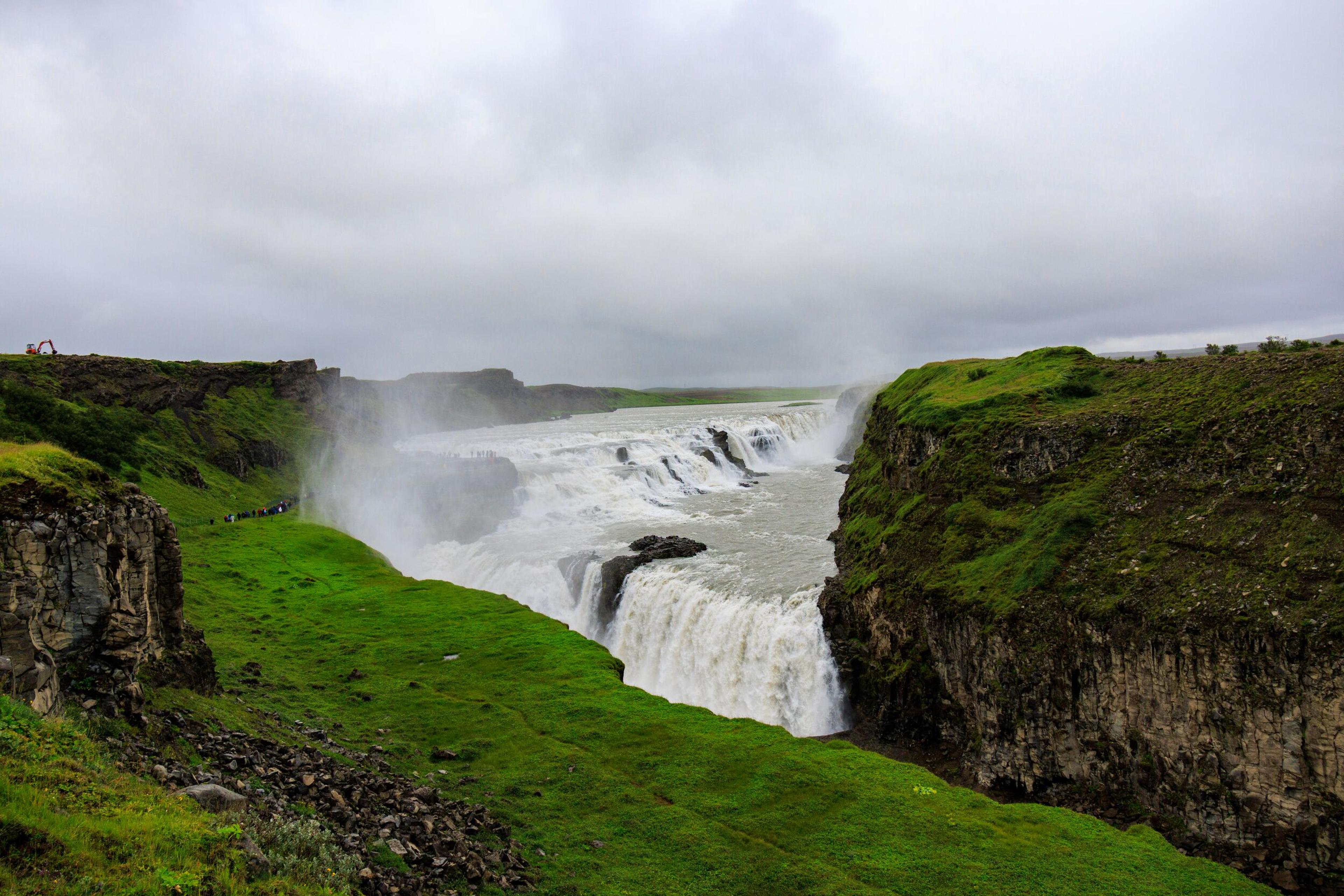 A wide, powerful waterfall cascades through a rocky gorge lined with green cliffs under a cloudy sky, with mist rising.