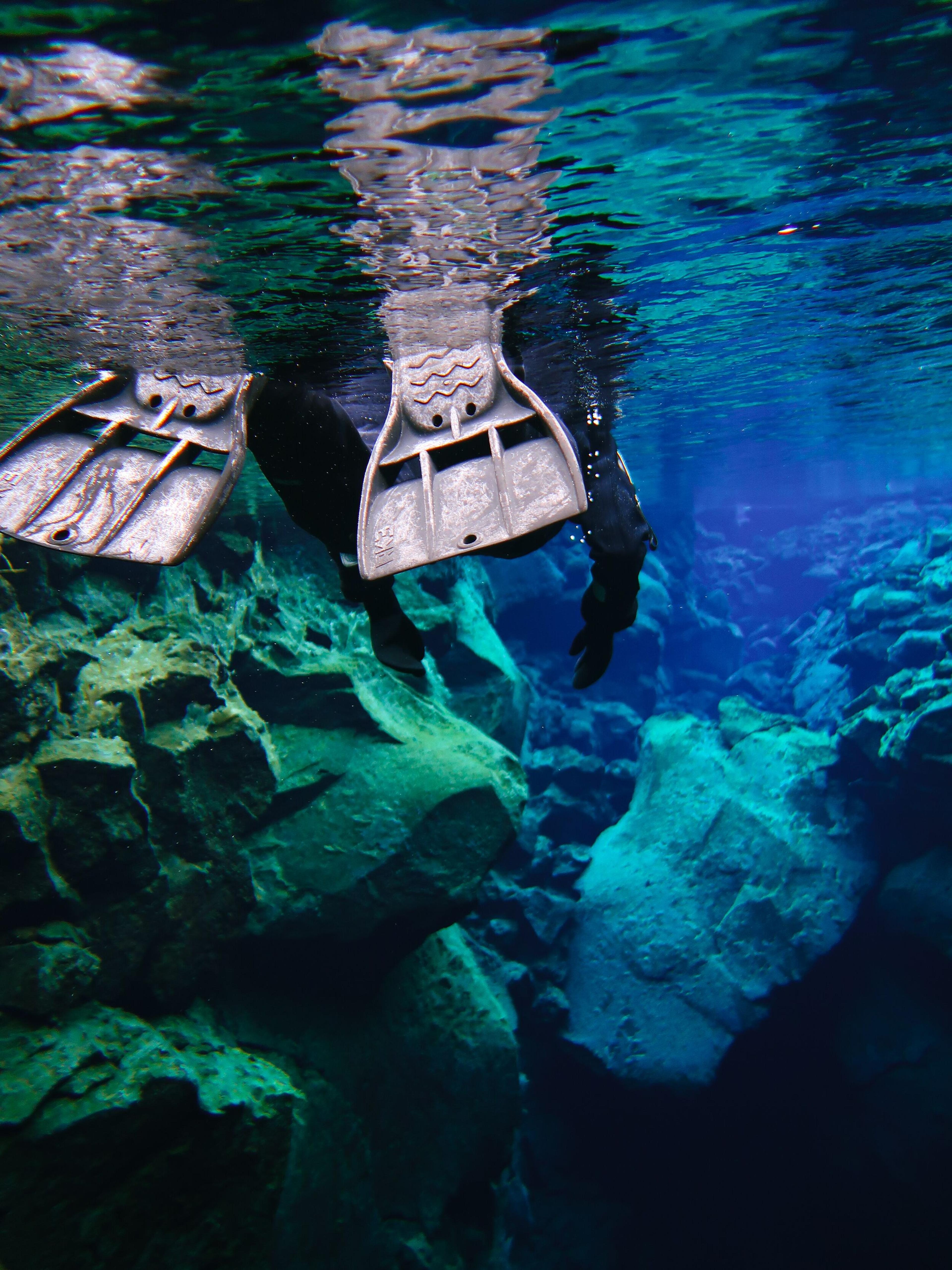 Snorkeler's fins visible in clear blue water above an underwater rocky rift.