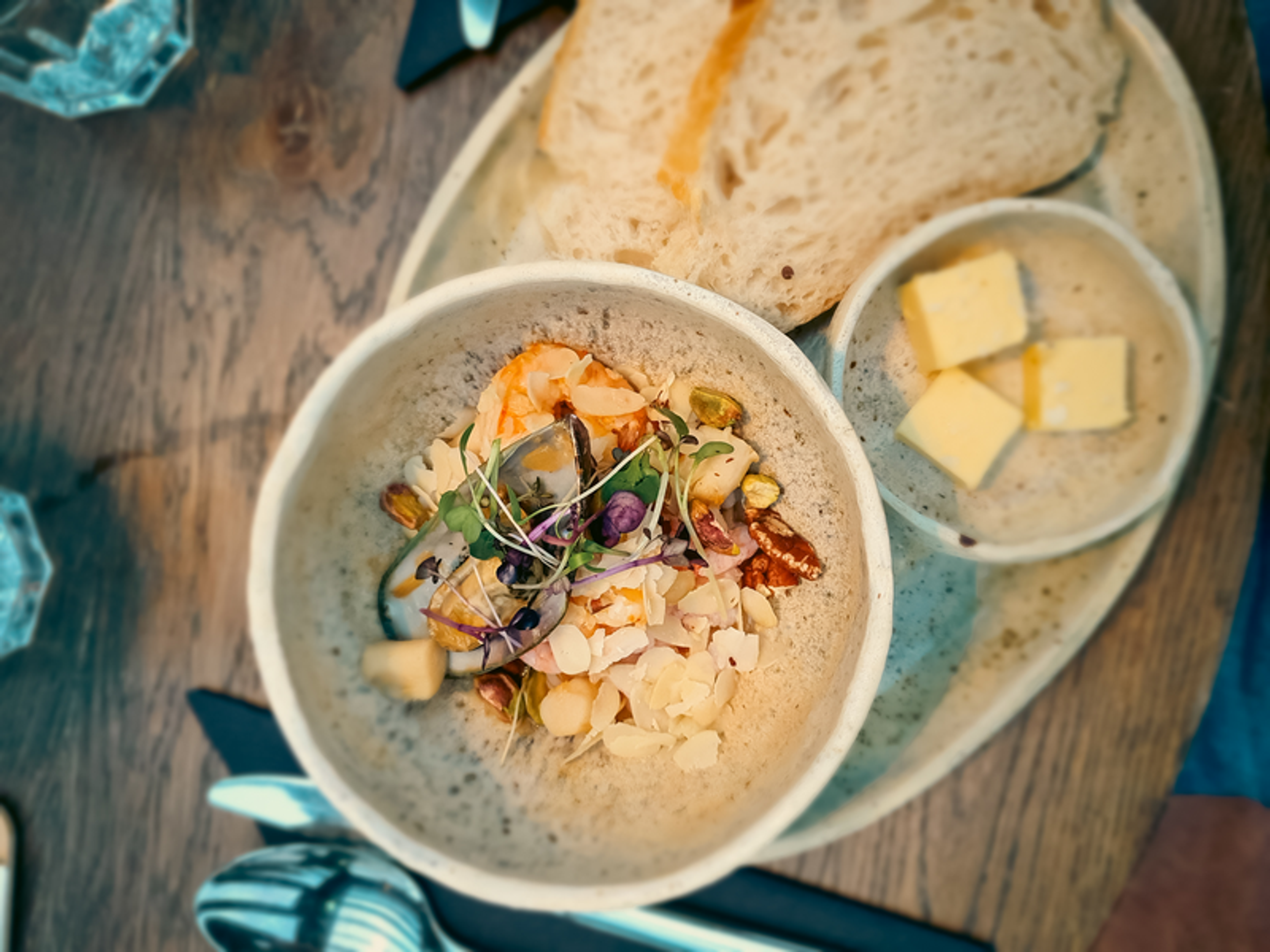 A bowl of creamy soup garnished with herbs and nuts, accompanied by slices of bread and butter on a wooden table.