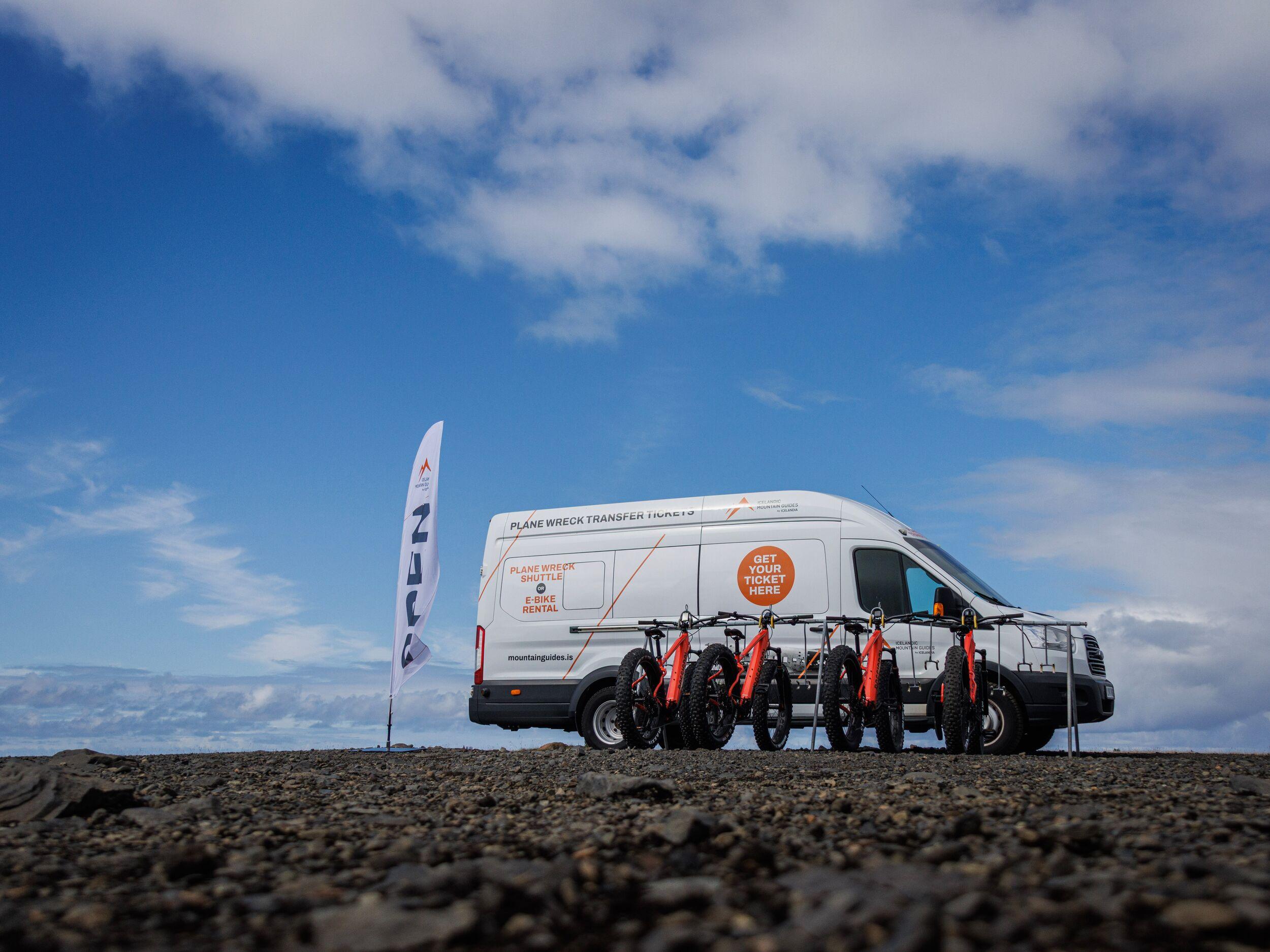 White van with multiple orange fat-tire e-bikes on a dark, rocky landscape under a blue sky, advertising "Plane Wreck Transfer" and "E-Bike Rental."