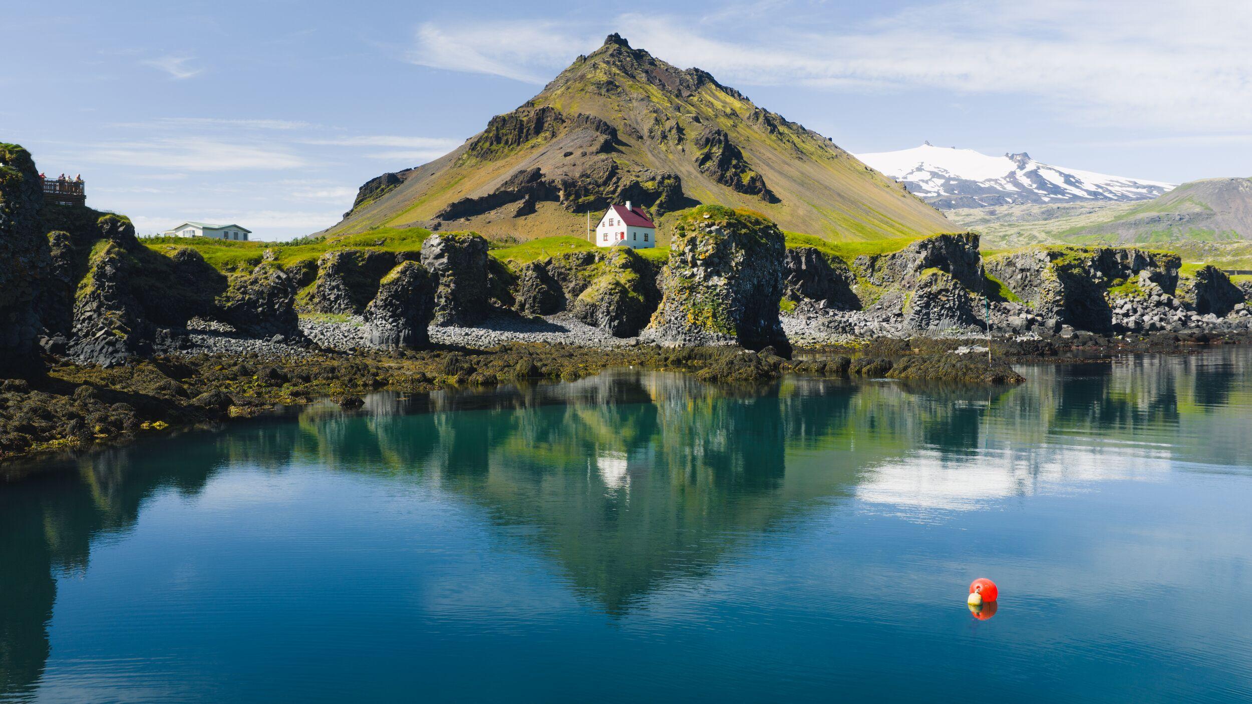 White house with red roof on a green clifftop overlooking calm water, flanked by rugged cliffs and a misty mountain.