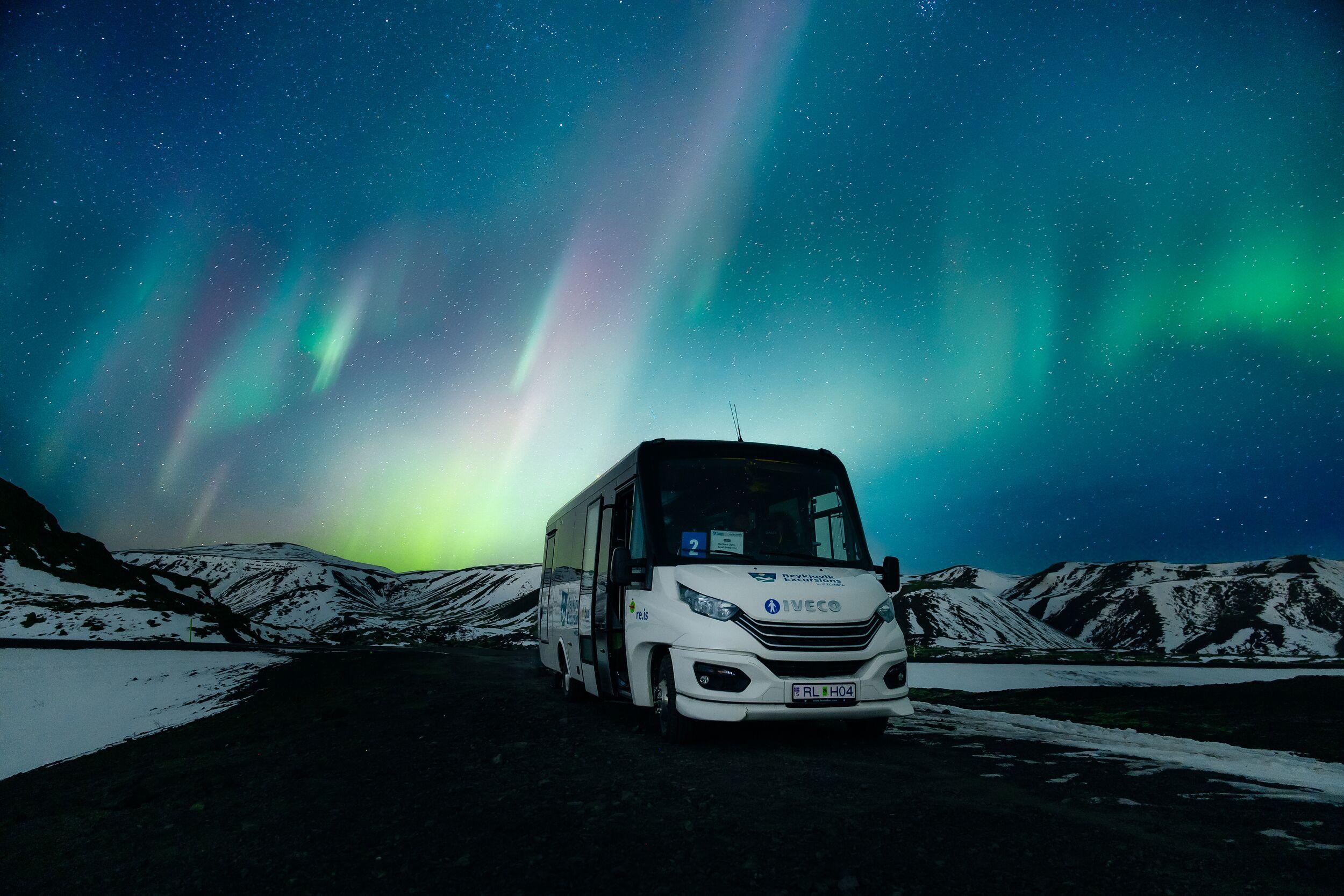 A white minibus parked on a road in a snowy mountain landscape under a vibrant green and purple aurora.