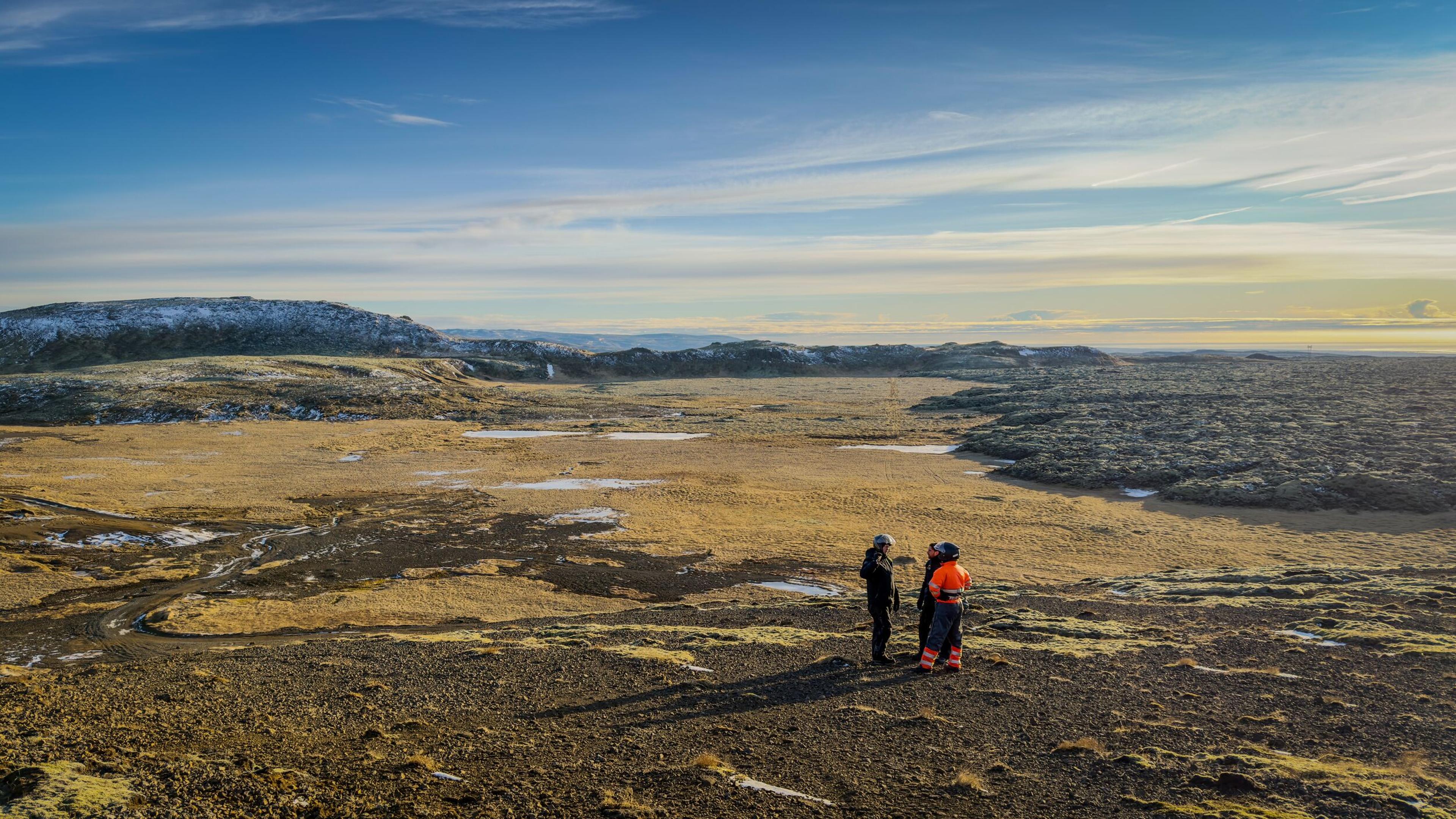 Two people stand on rocky ground, overlooking a vast landscape of golden grassland, dark lava flows, and distant hills under a bright sky.