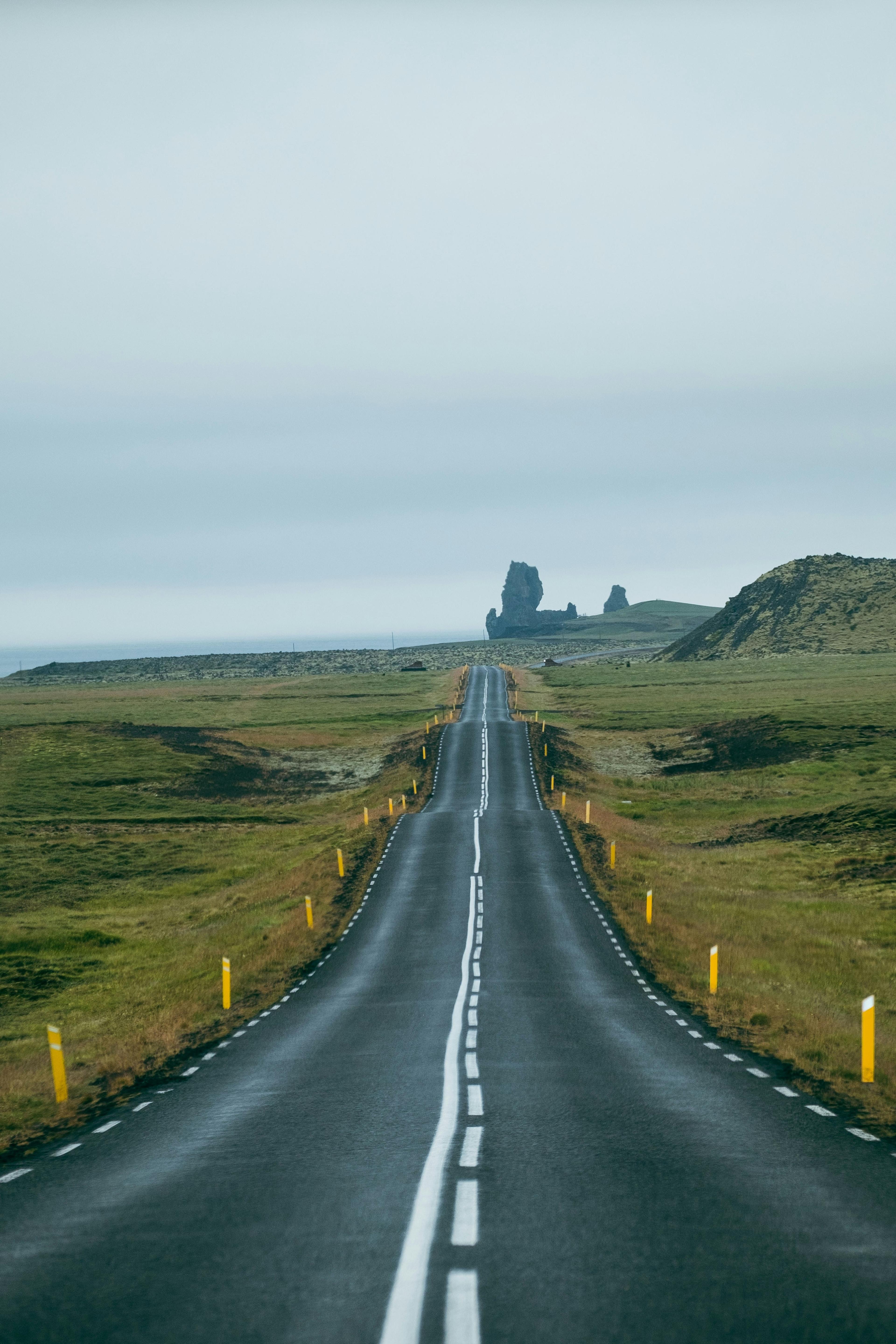 A long, straight road stretches through the vast Icelandic landscape, flanked by green fields and leading toward distant rugged rock formations under an overcast sky.