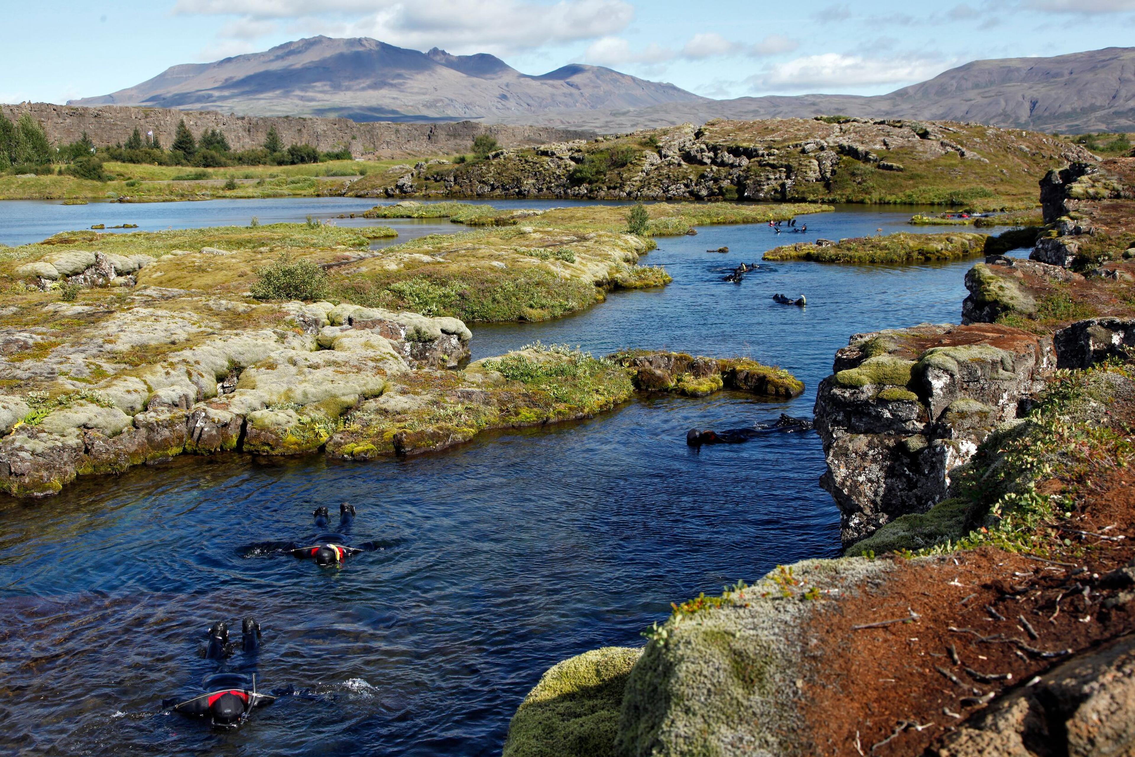 Scuba divers in a serene, clear water river surrounded by moss-covered lava rocks and a lush green landscape, with a backdrop of distant mountains under a blue sky.