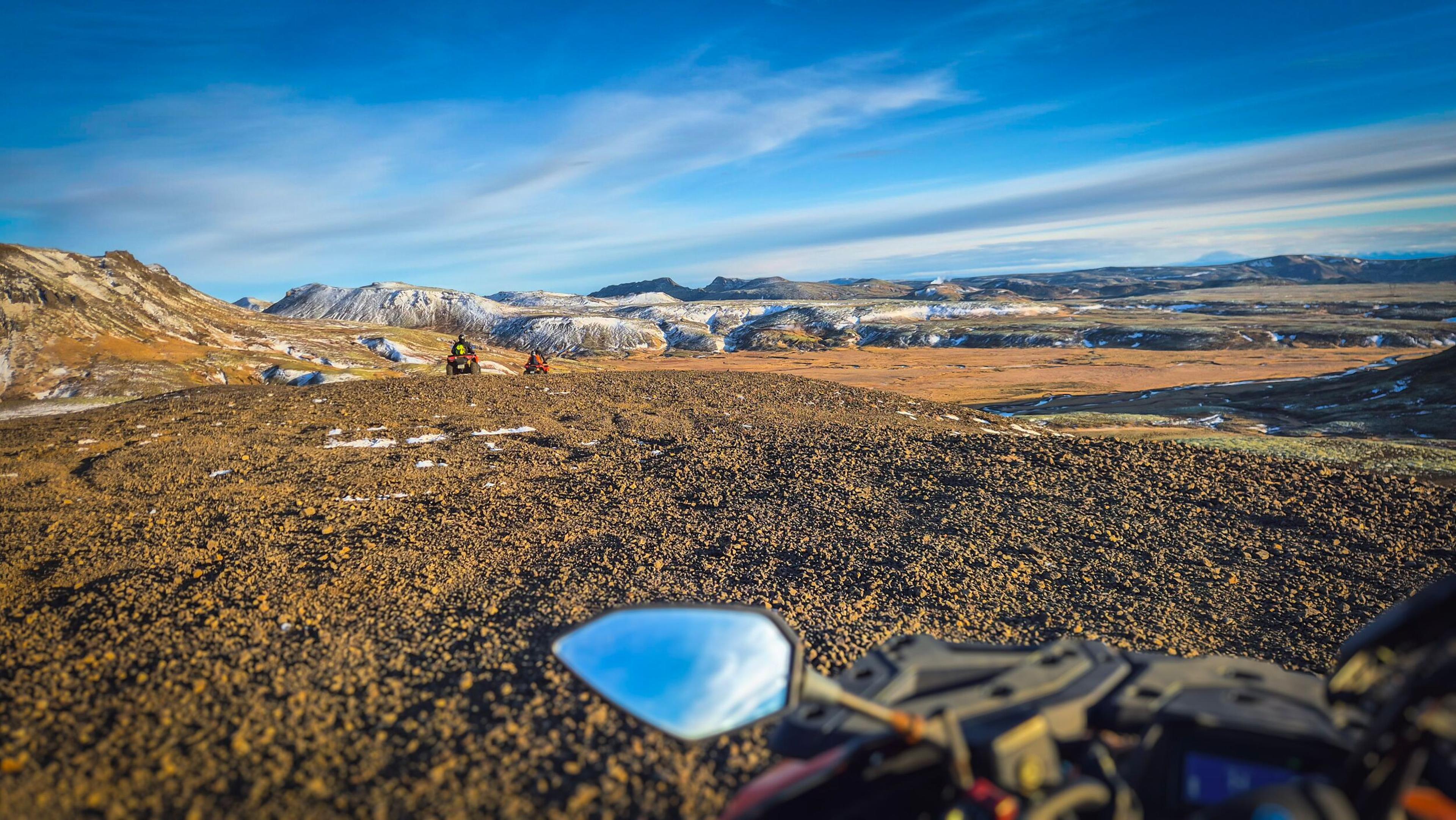 First-person view from an ATV on a rugged, snow-dusted landscape with distant mountains and other riders.