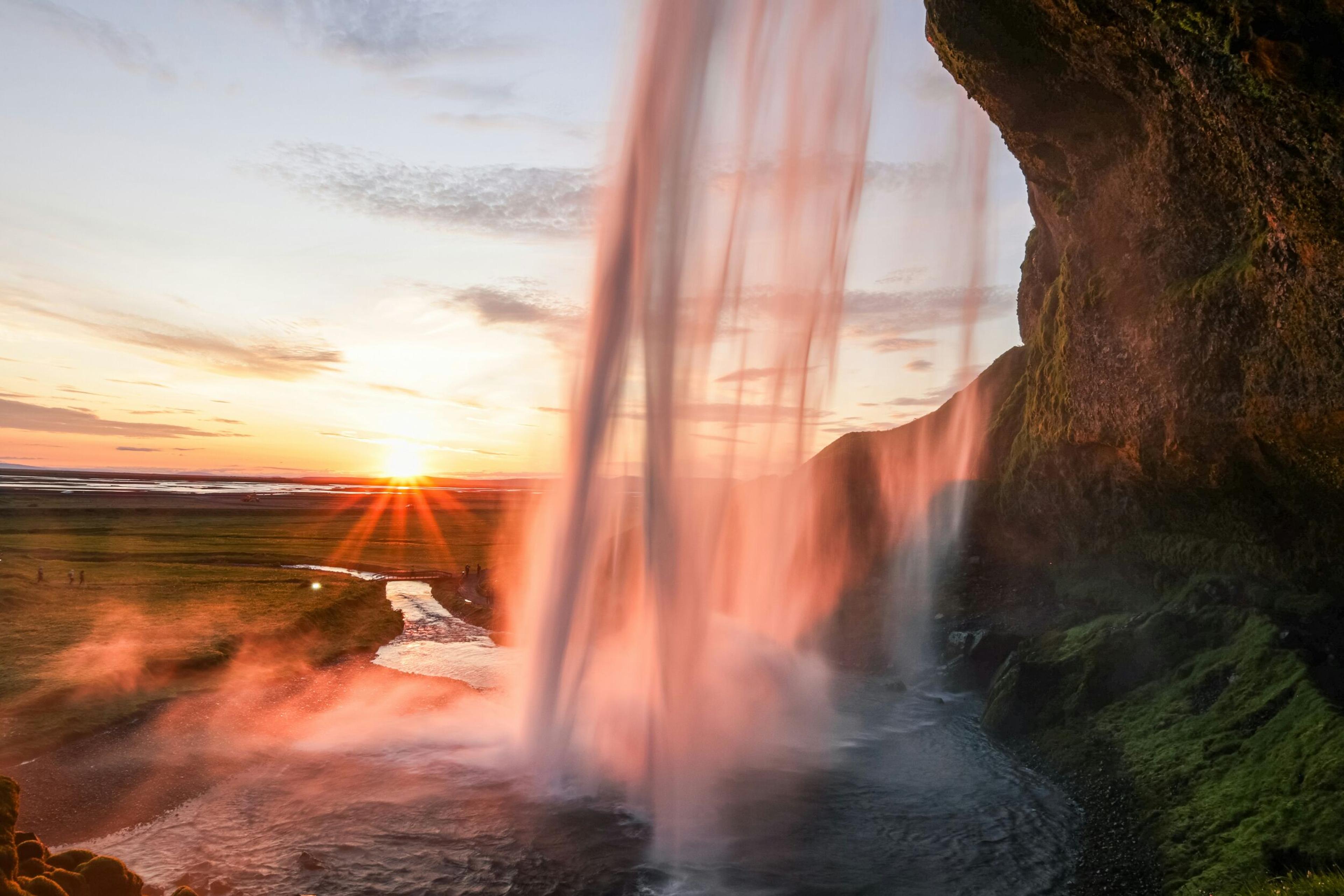 Sunset view from behind Seljalandsfoss waterfall in Iceland, with warm light casting a glow on the water and surrounding landscape.