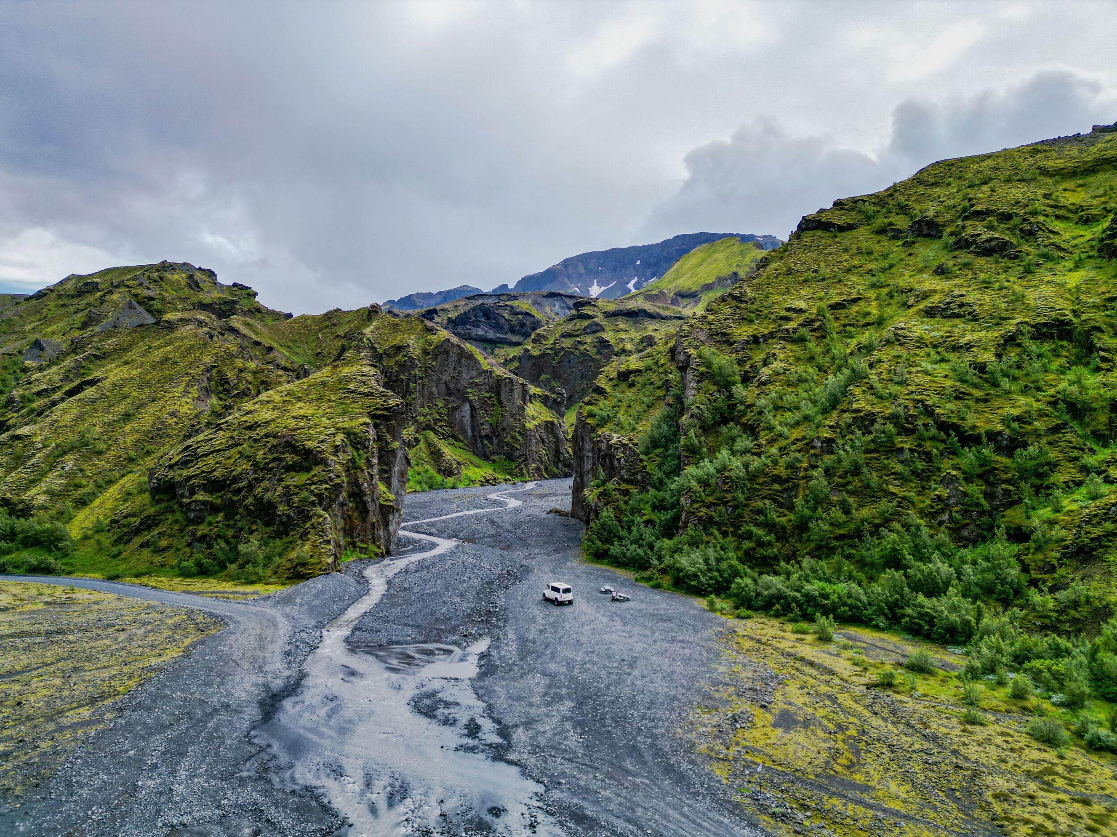 A white camper van on a winding rocky riverbed through a lush, mossy green valley under a cloudy sky.