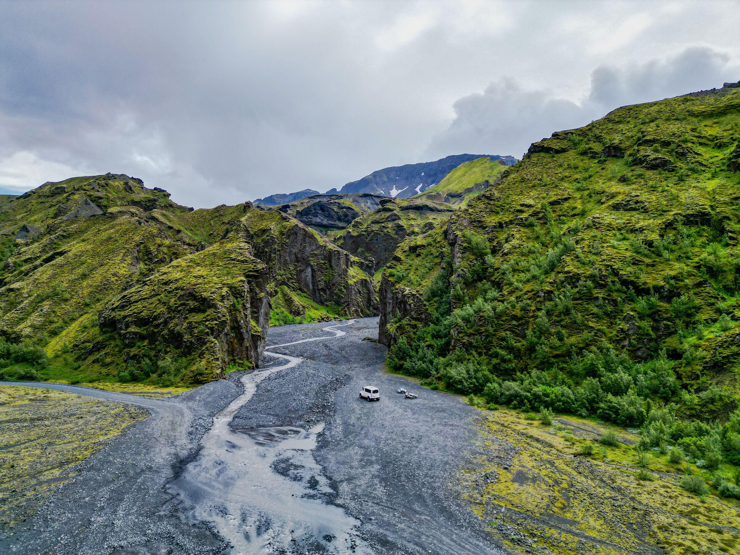 A white camper van on a winding rocky riverbed through a lush, mossy green valley under a cloudy sky.