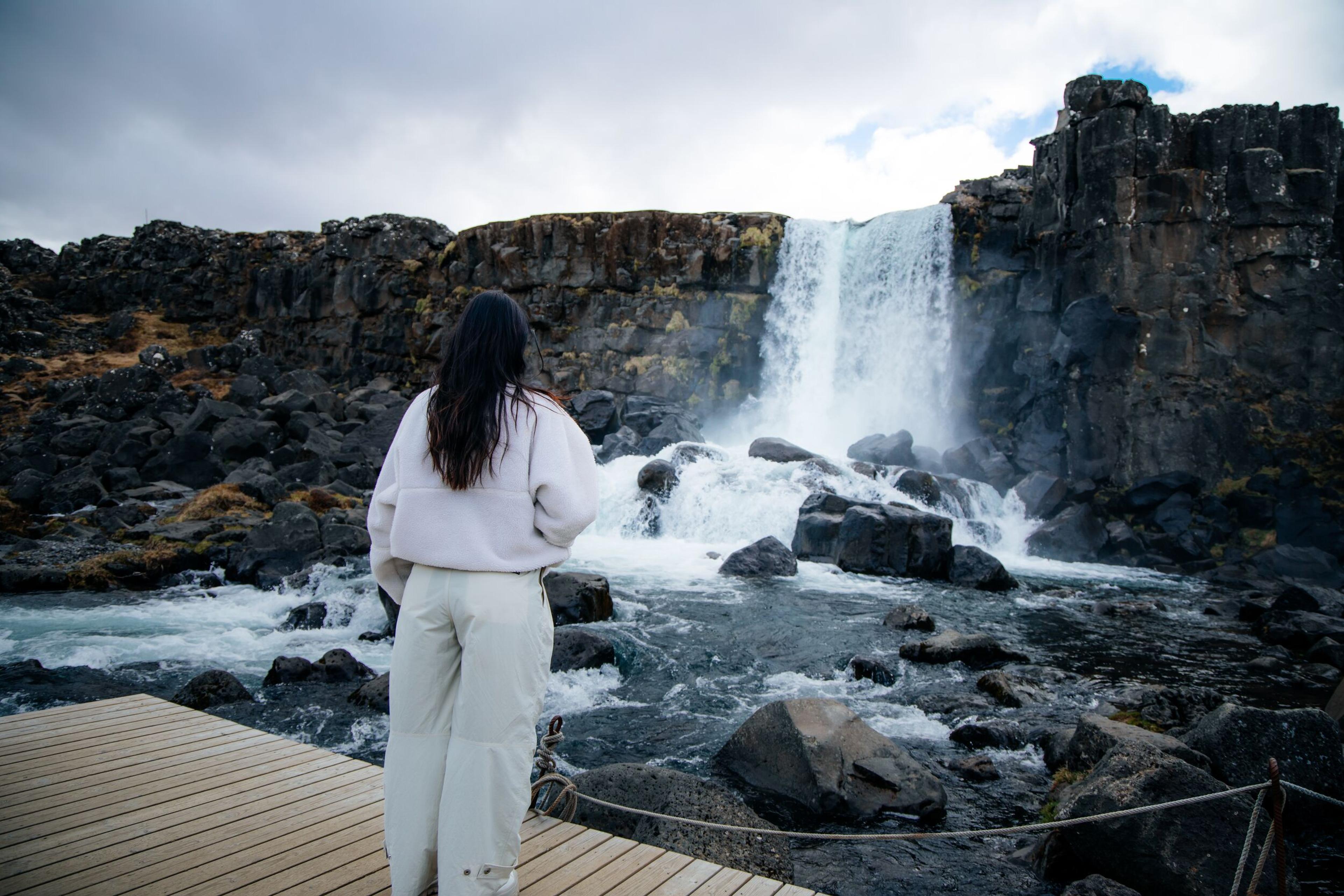 A person stands on a wooden platform, looking at a waterfall cascading over dark rocky cliffs.