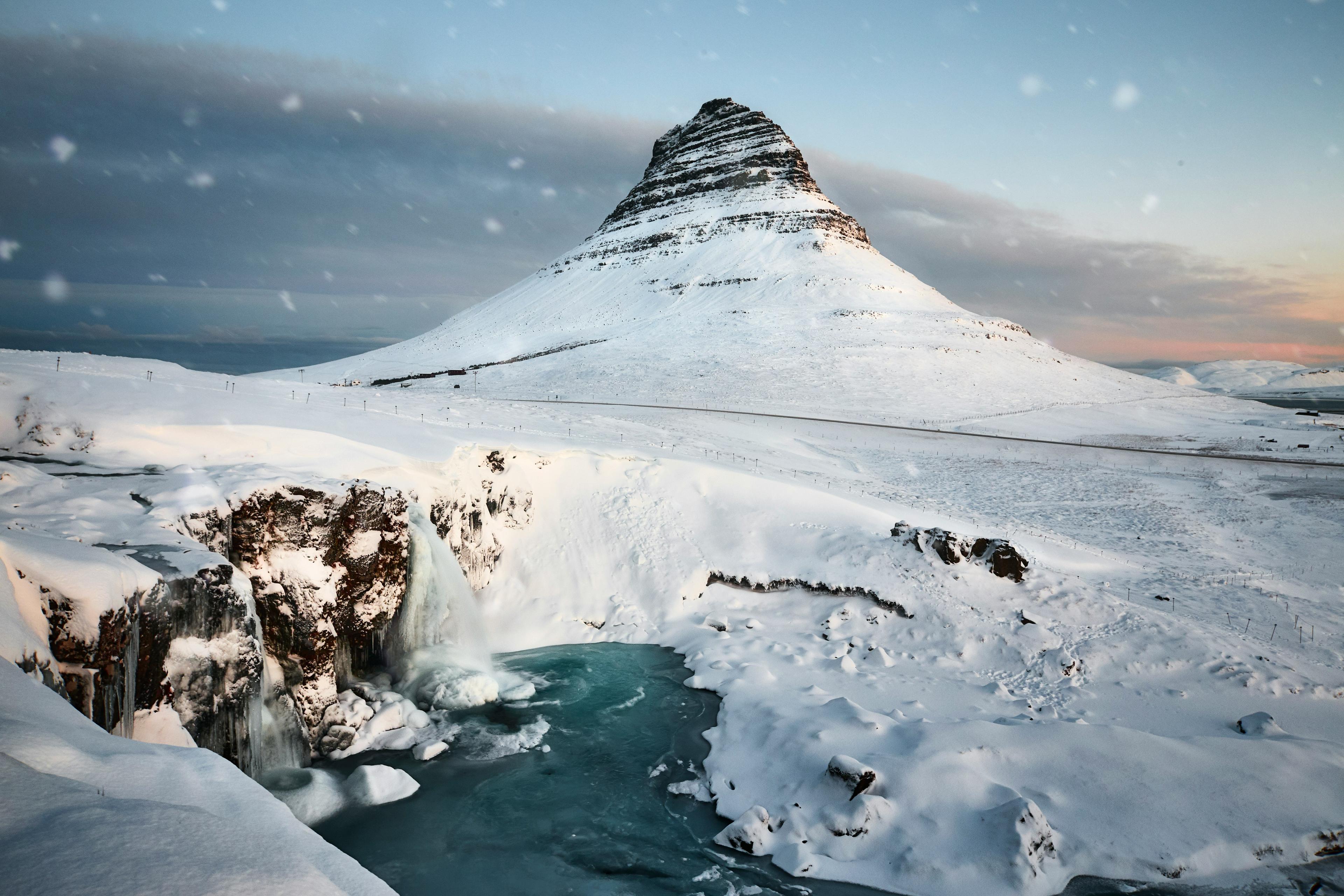 Snow-covered Kirkjufell Mountain with a frozen waterfall and icy blue waters in the foreground, showcasing a serene winter landscape.