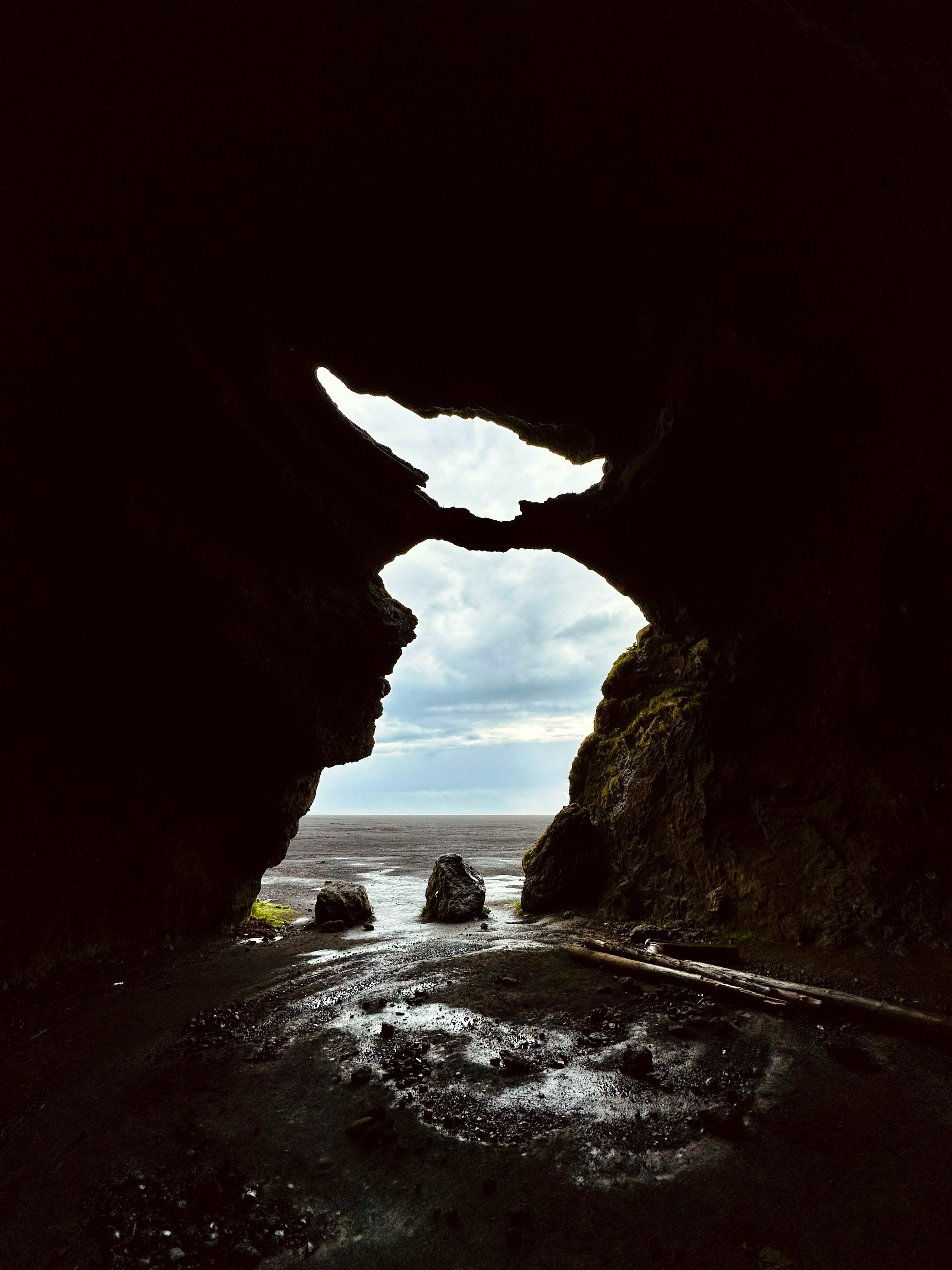 View from a dark cave showing a wet beach, ocean, and cloudy sky through two rock openings.
