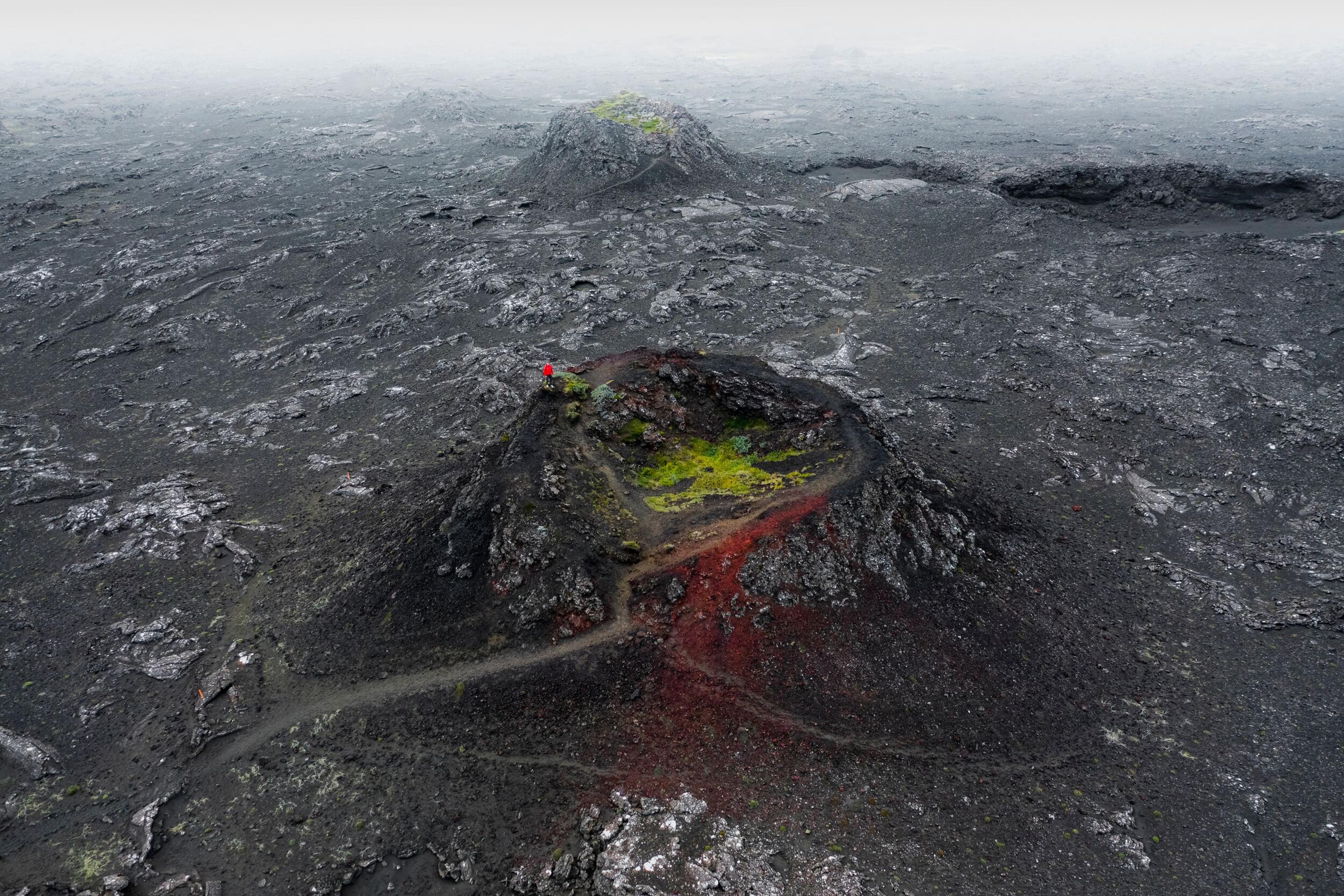 Aerial view of a volcanic landscape with a small crater, featuring red and green patches of vegetation amidst dark lava fields.