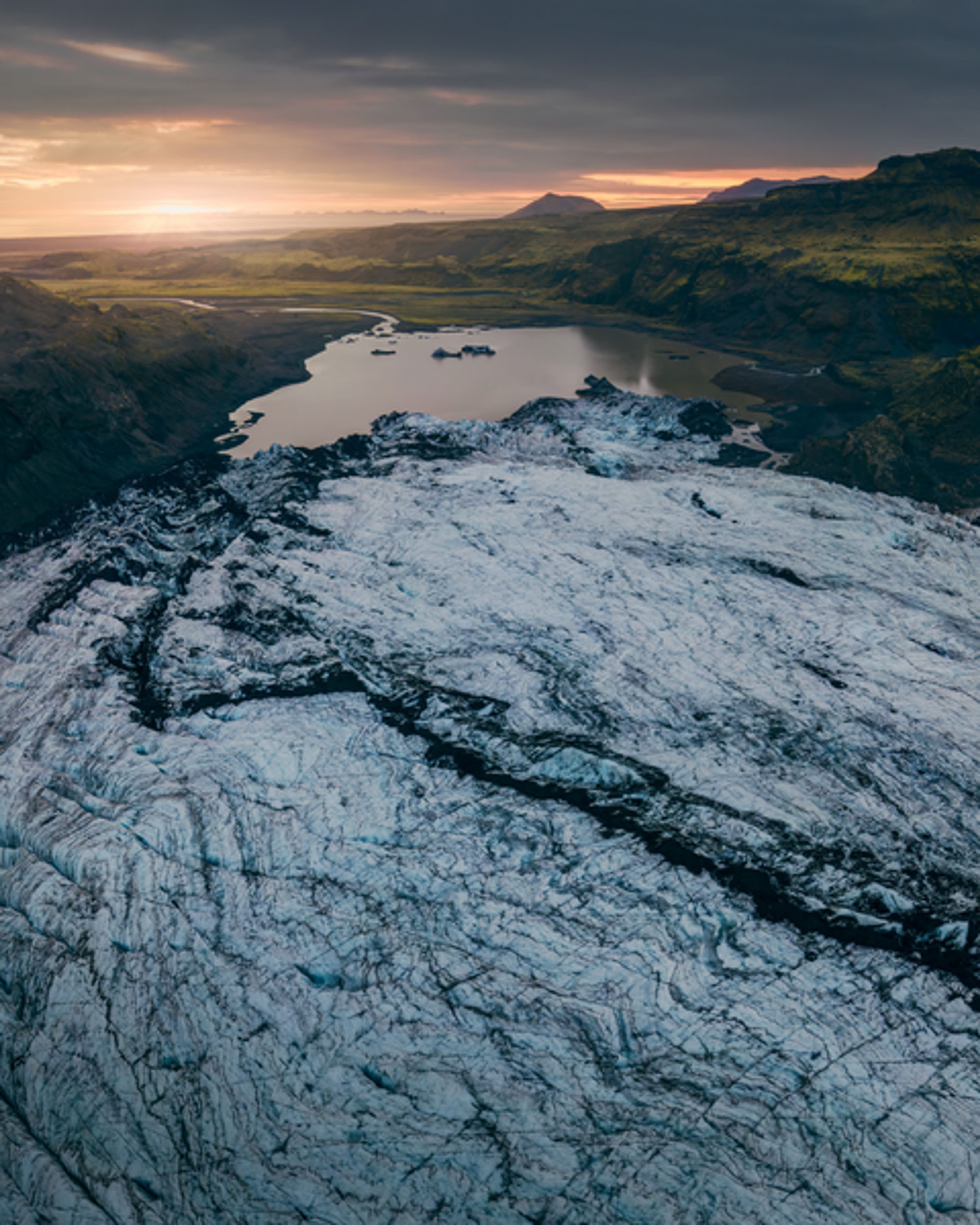 Aerial view of Mýrdalsjökull glacier with ice formations, surrounded by a vast landscape and a lake, under a sunset sky.