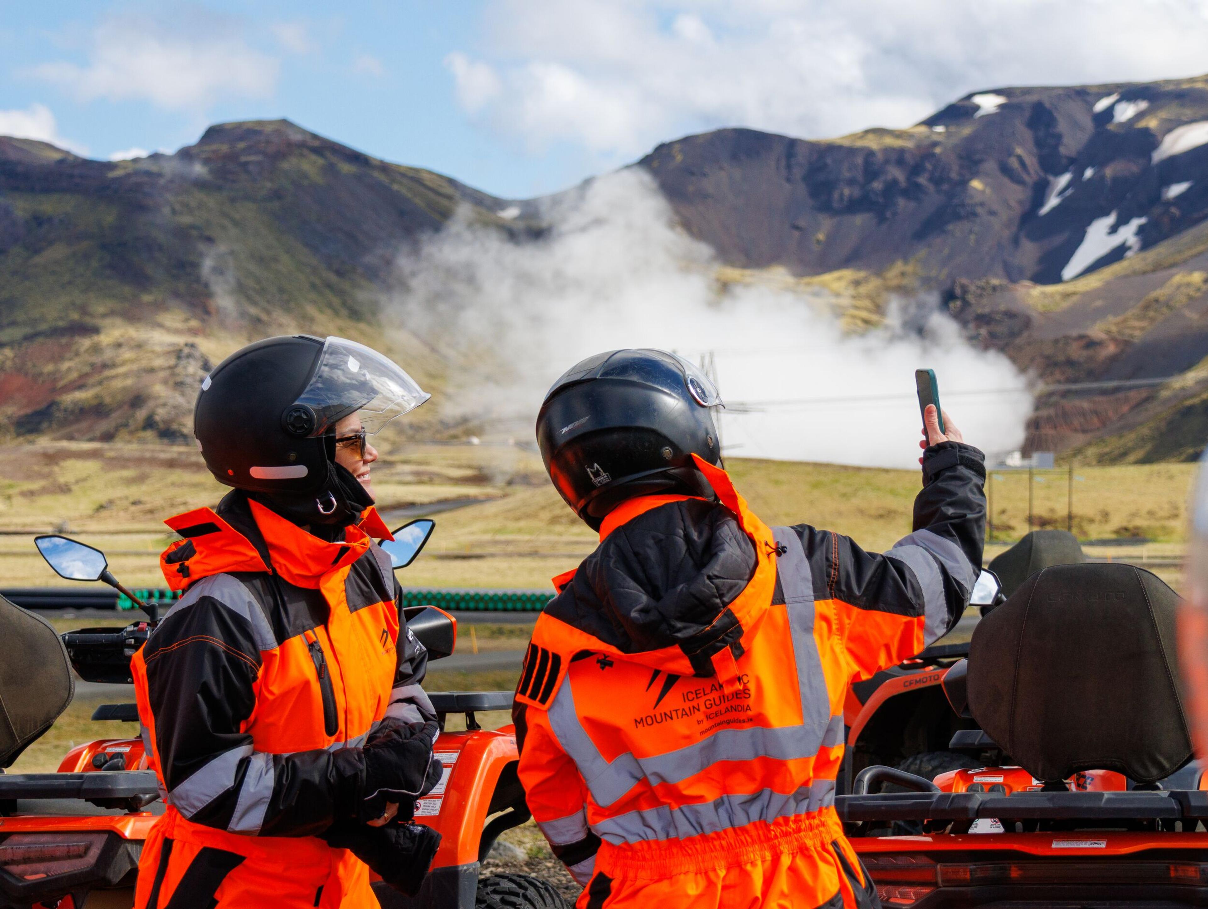 Two ATV riders in orange suits and helmets, one taking a photo of a steaming geothermal landscape.