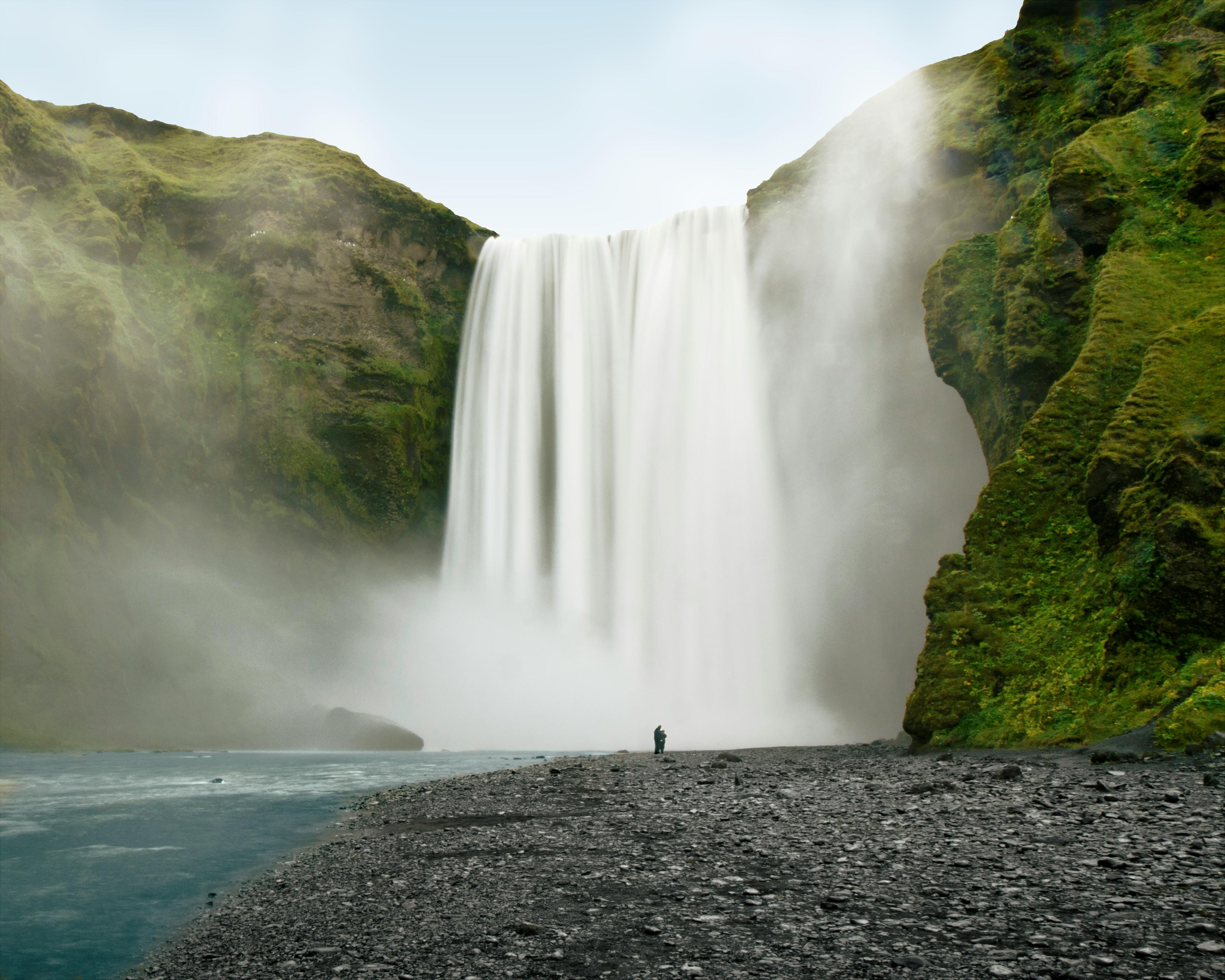 A solitary figure stands at a distance, dwarfed by the immense scale of a powerful waterfall plunging into a misty pool, surrounded by lush green cliffs.