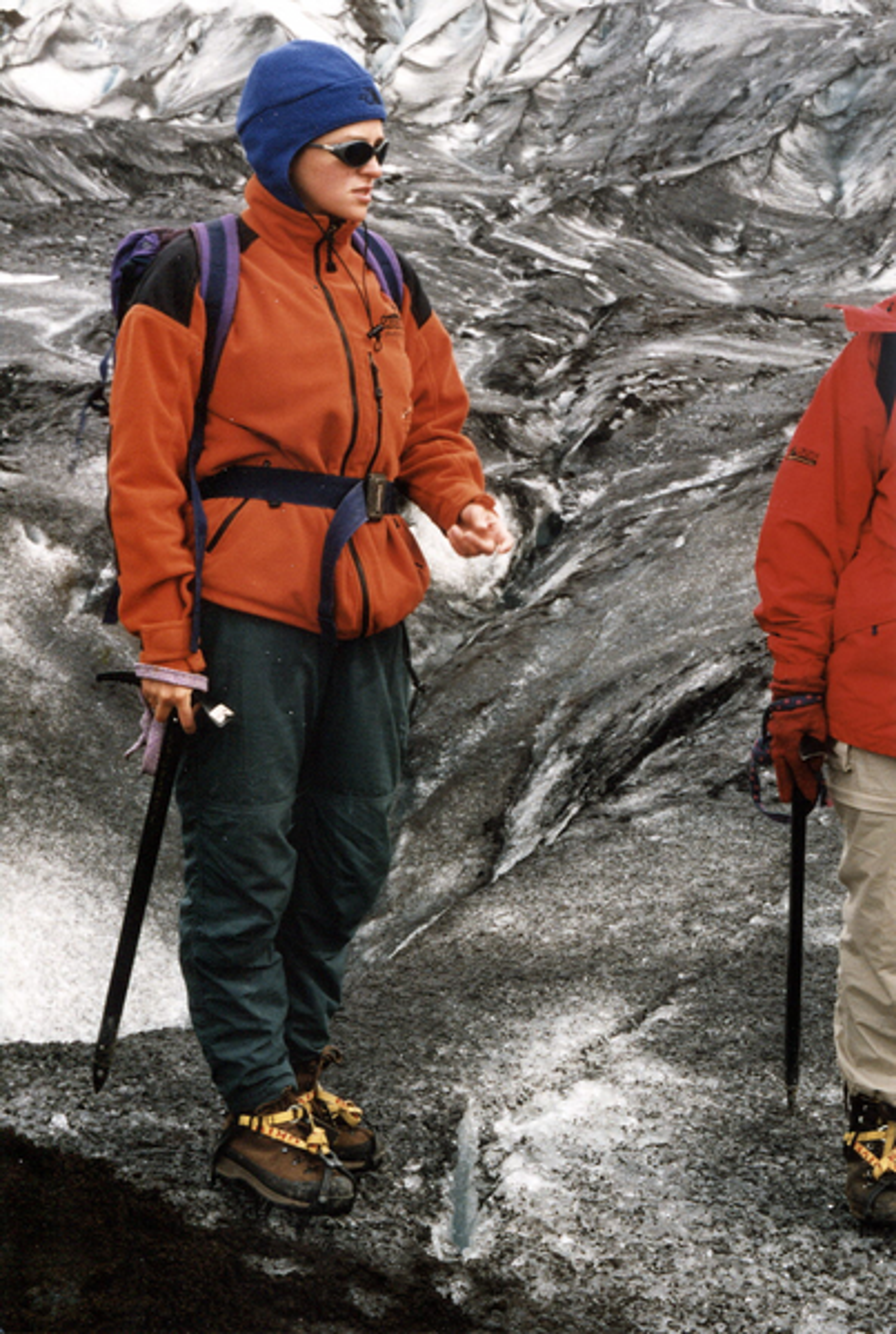 A young woman outfitted in cold weather hiking gear stands on a rocky terrain with a glacier in the background.