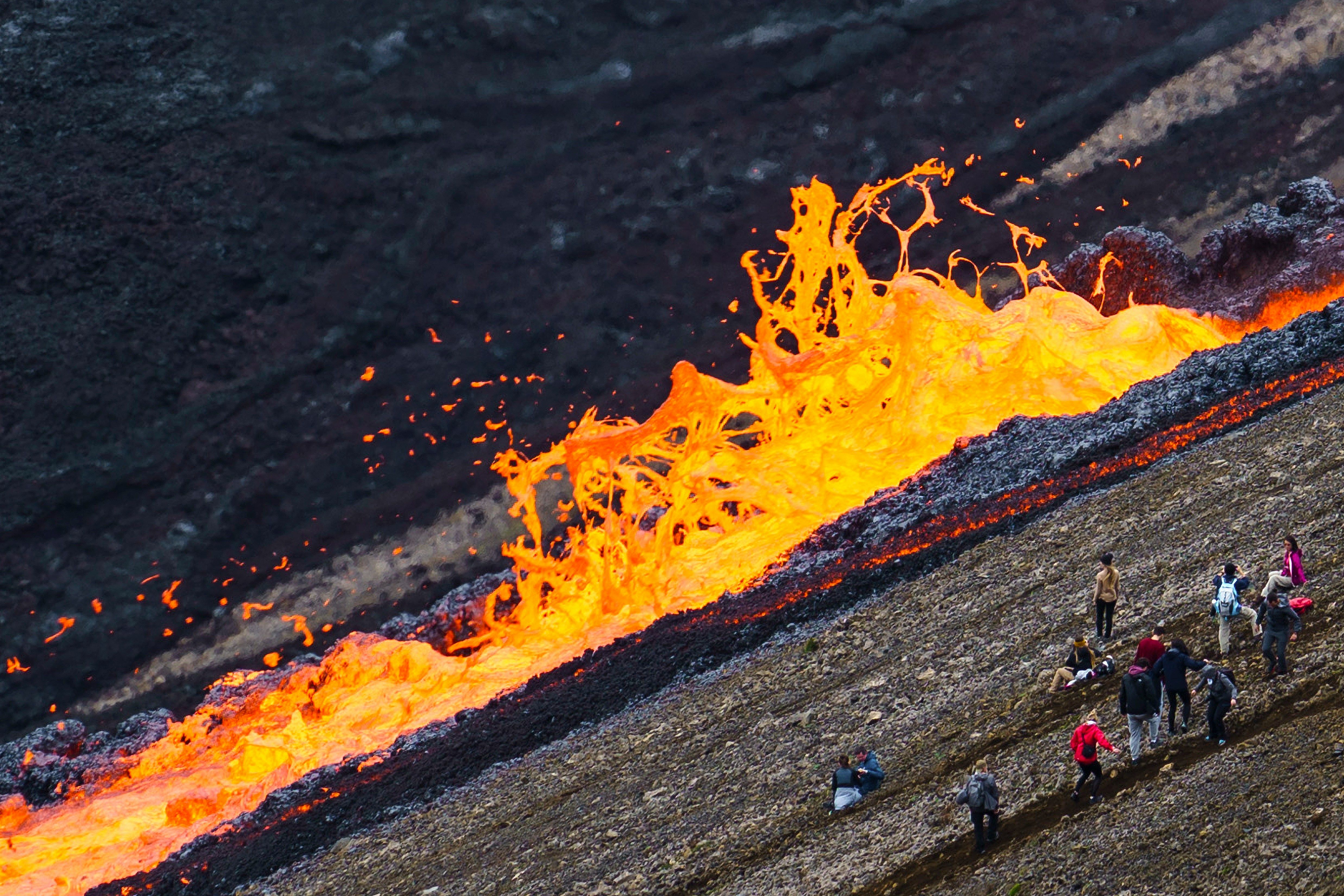 "A wide view of a lava flow cutting through a valley in Iceland, with a group of people gathered near its edge. 