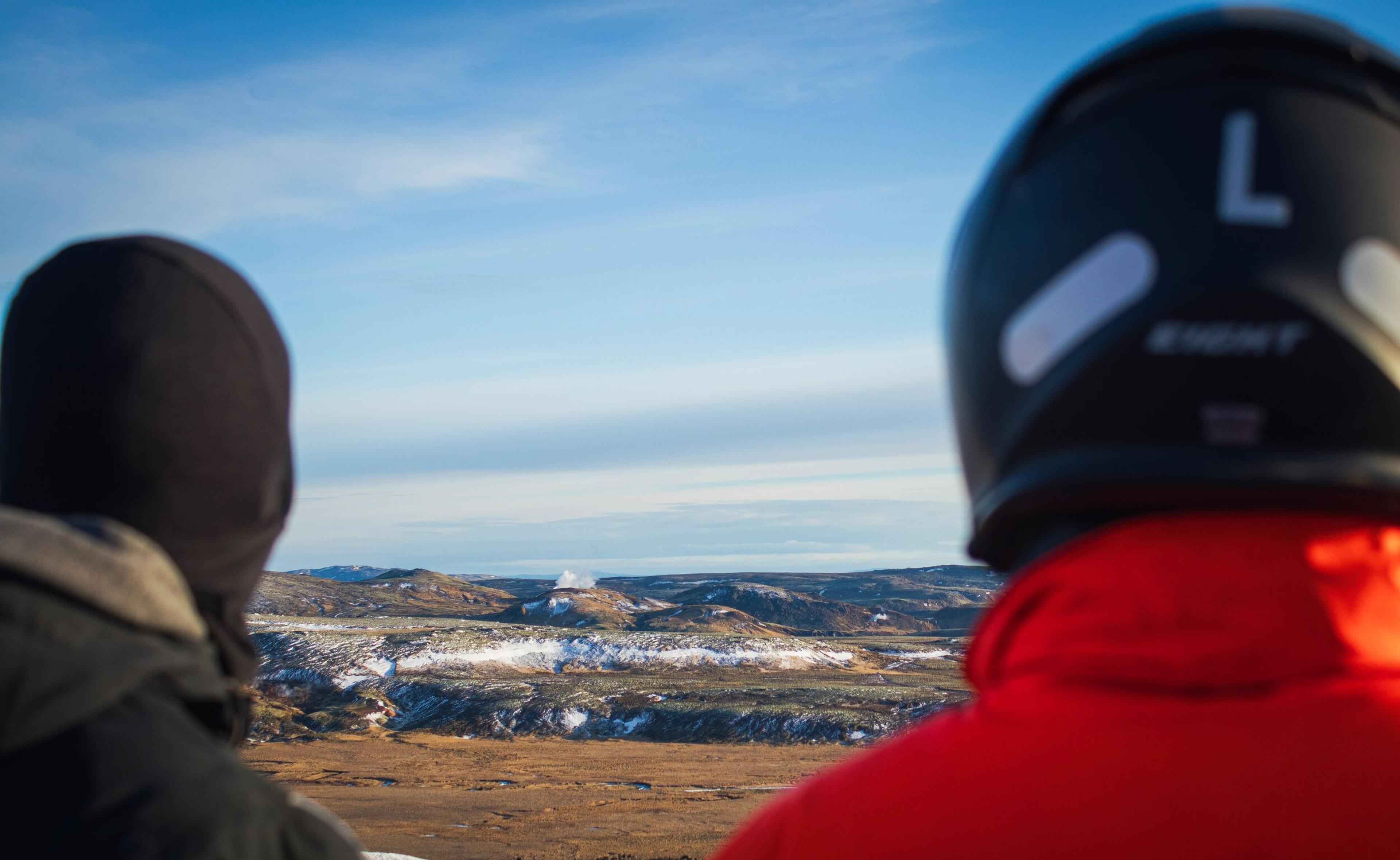 Two people, one wearing a black helmet, gaze at a vast, snowy landscape with steam rising from distant hills under a blue sky.