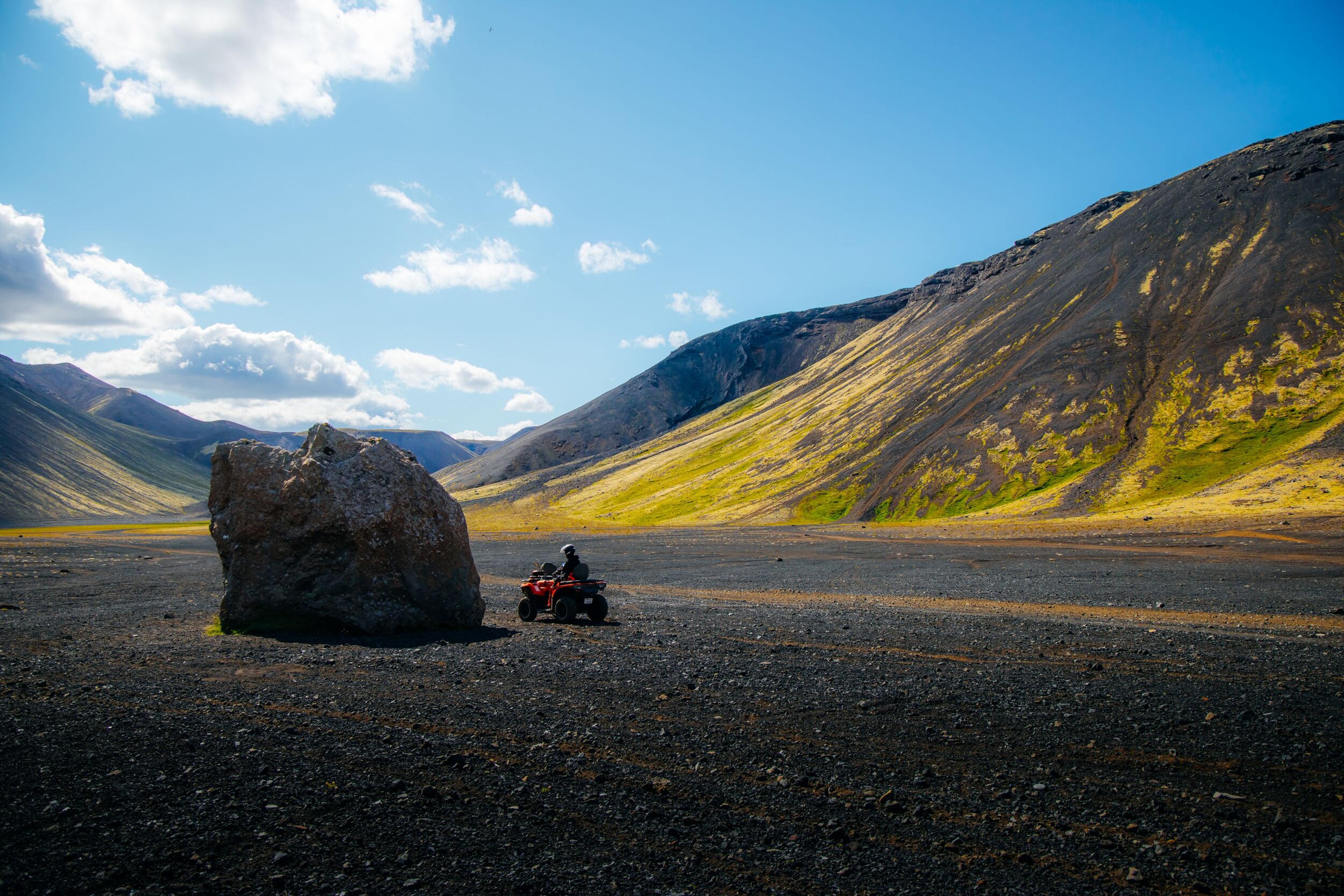 A person on an ATV next to a large boulder in a vast, dark valley with mountains and a blue sky.