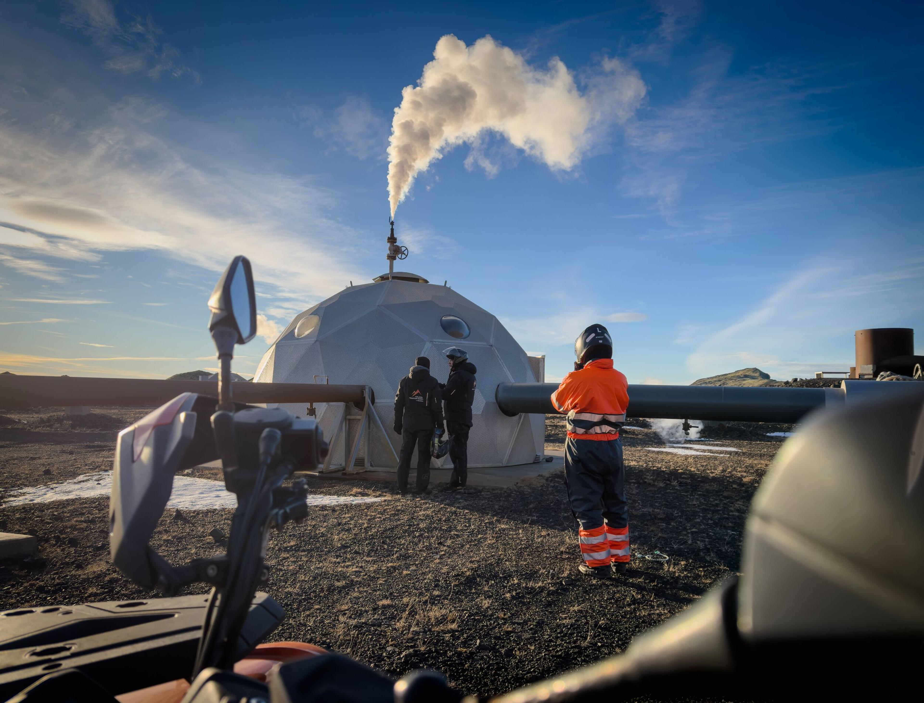 Geodesic dome facility emitting steam, with workers and an ATV in a barren landscape.
