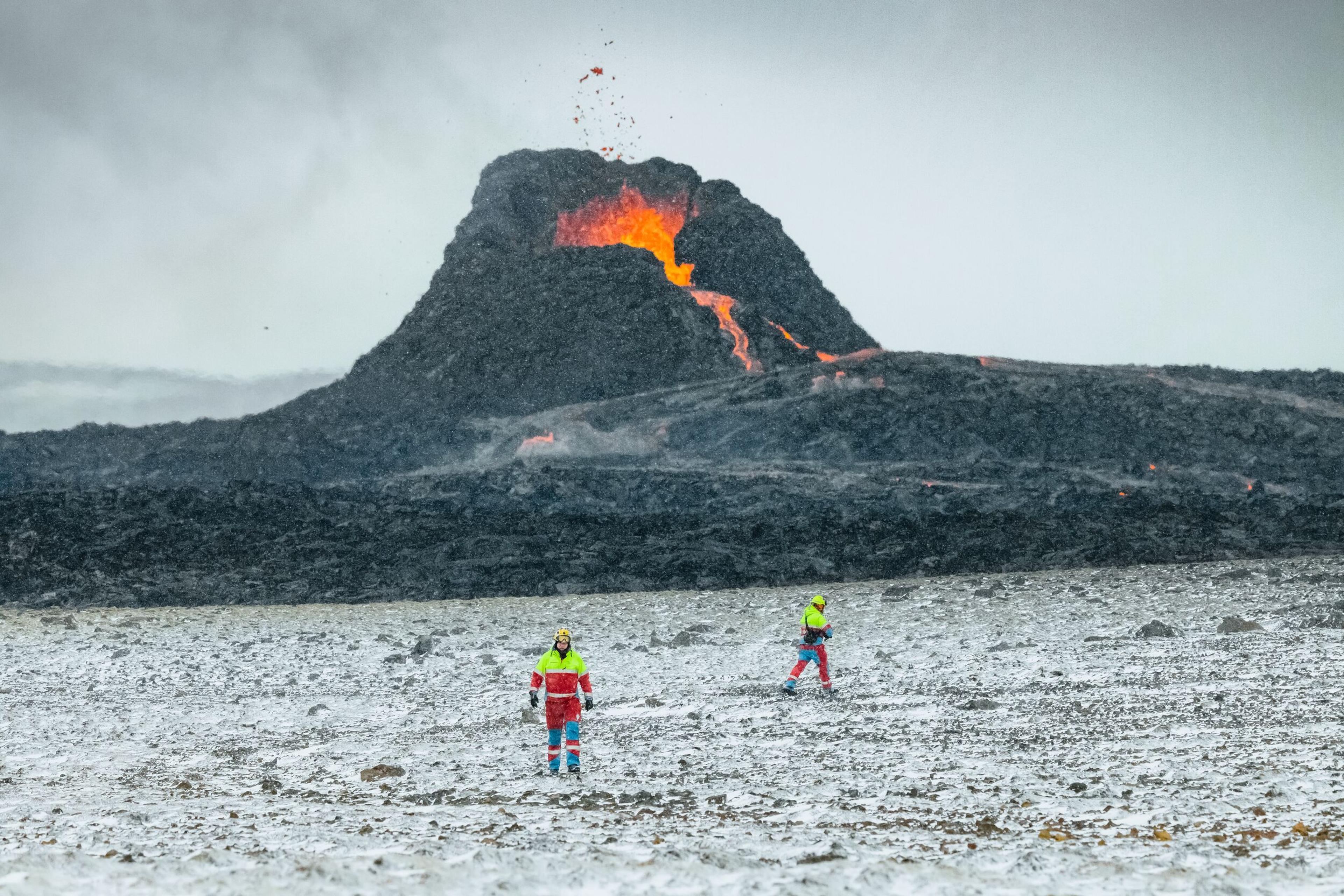 Two people in protective gear observe an active volcanic eruption, with molten lava flowing from a large, dark volcanic mound.