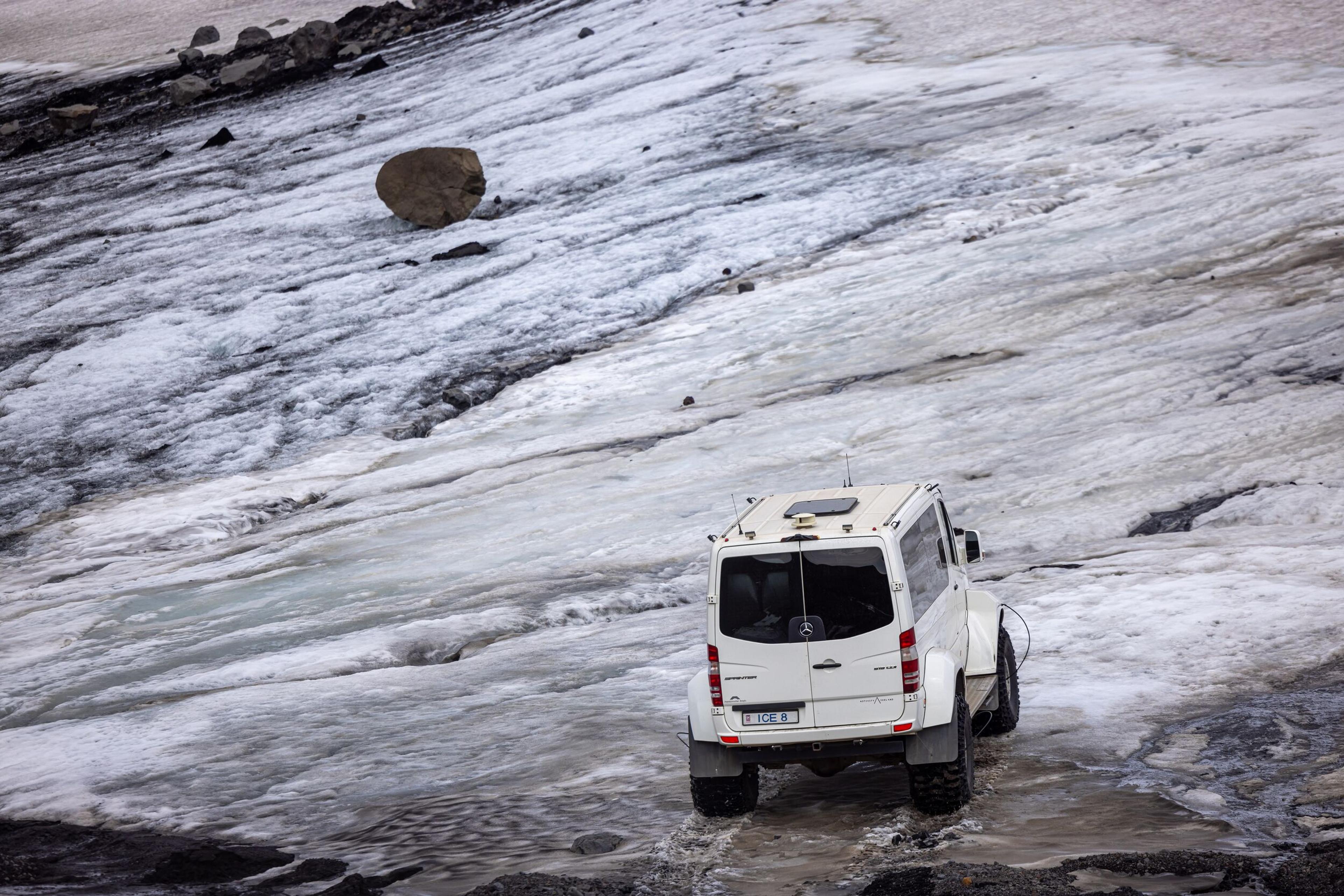  White off-road vehicle navigating across a glacier in Iceland, driving over icy terrain with cracks and patches of snow.