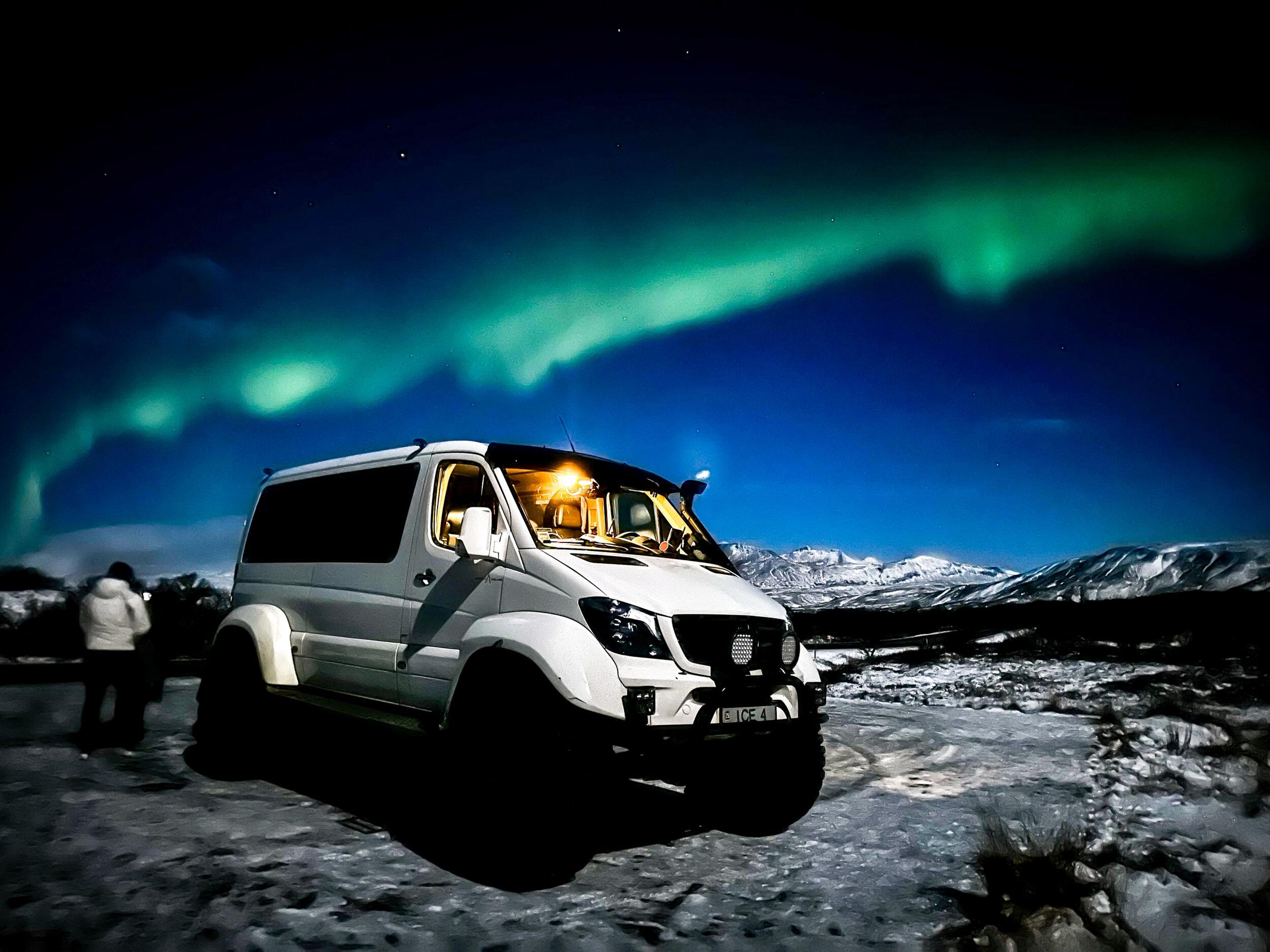 A white Icelandia super jeep with large tires parked in a snowy landscape under vibrant green Northern Lights.