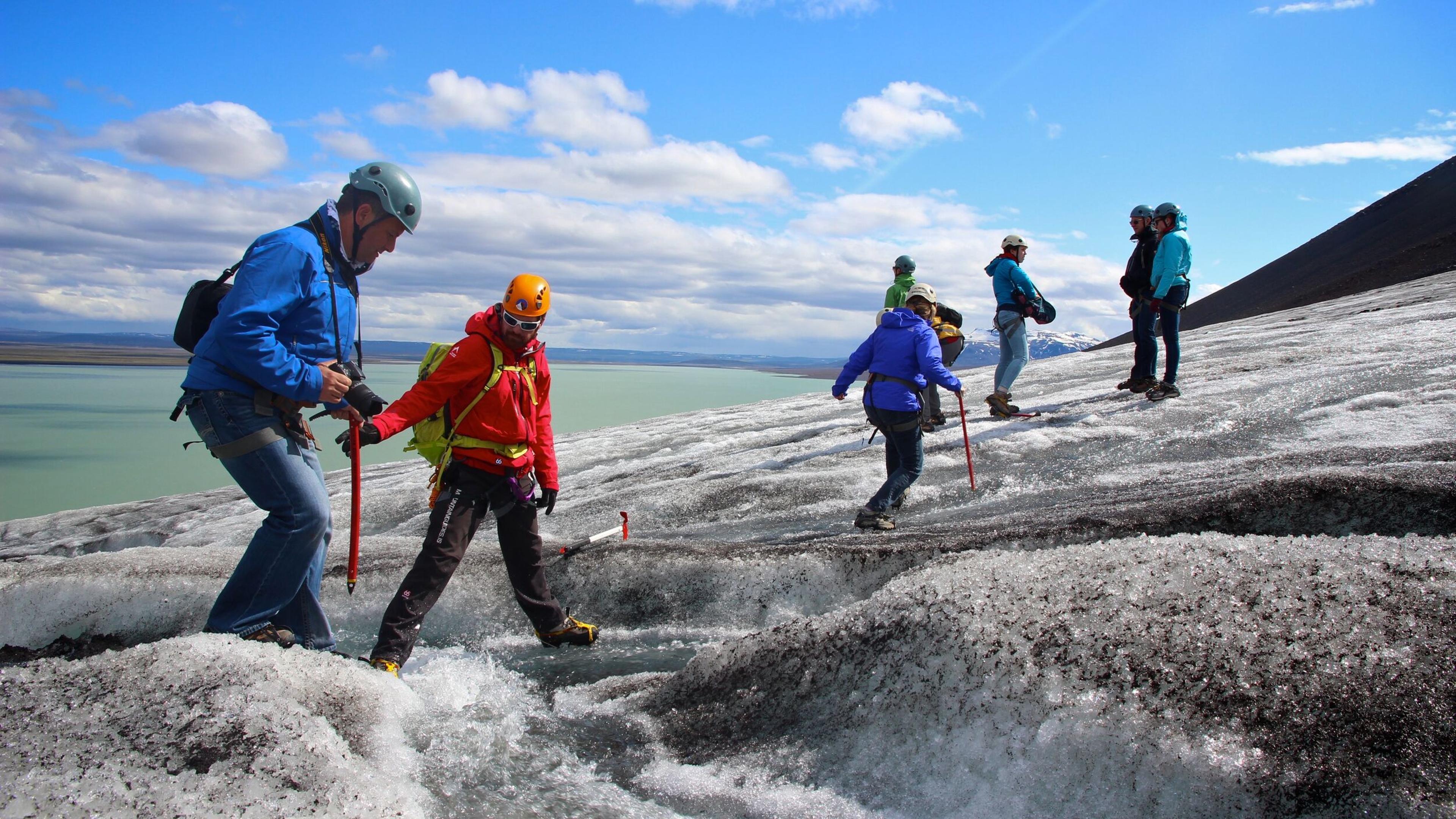 Hikers equipped with helmets and ice axes carefully traverse the icy terrain of Sólheimajökull glacier under a bright blue sky. A guided Sólheimajökull glacier hike offering adventure and stunning views in Iceland.