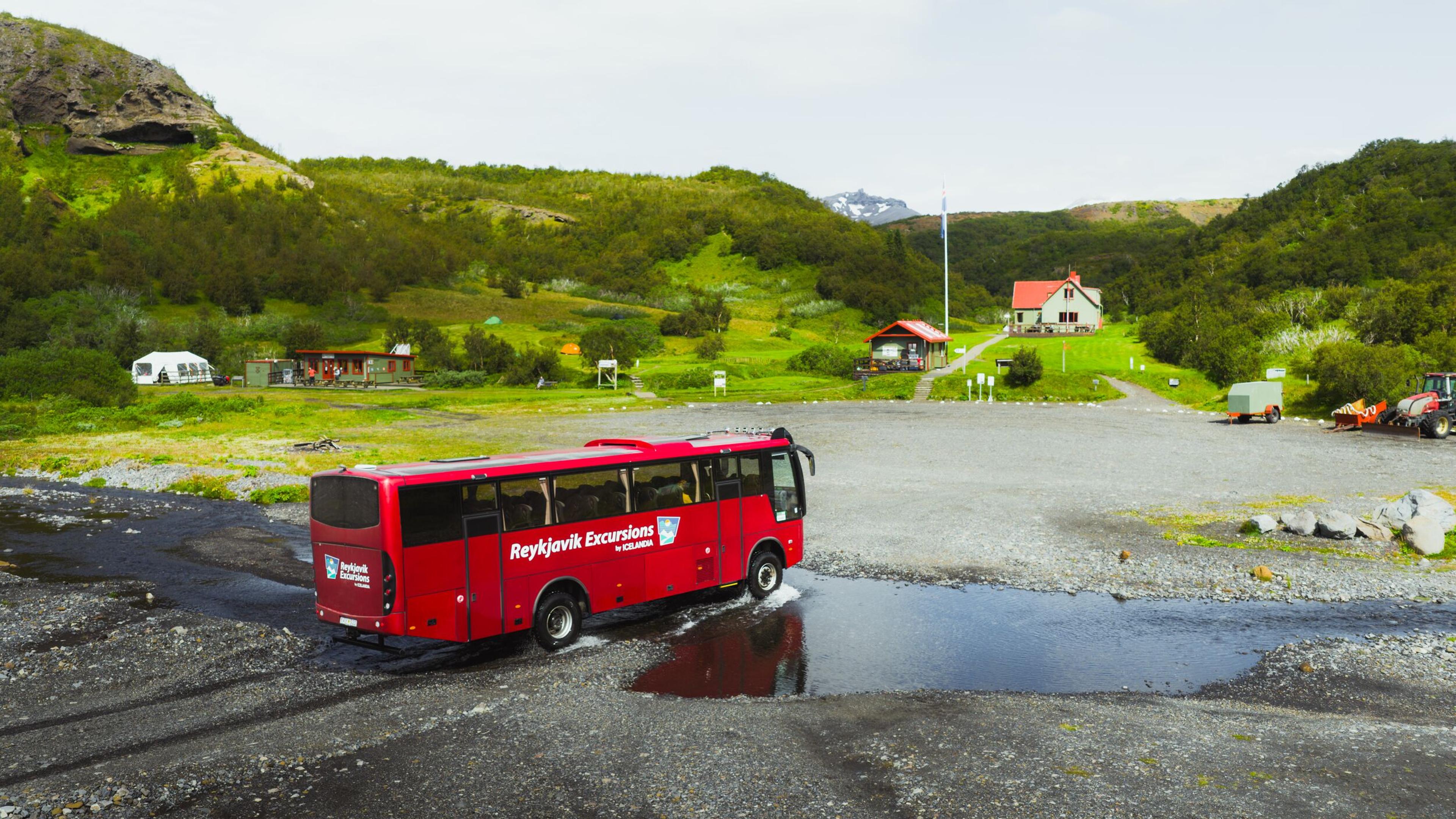 A red "Reykjavík Excursions" bus drives through a puddle on a gravel road in a green, mountainous landscape.