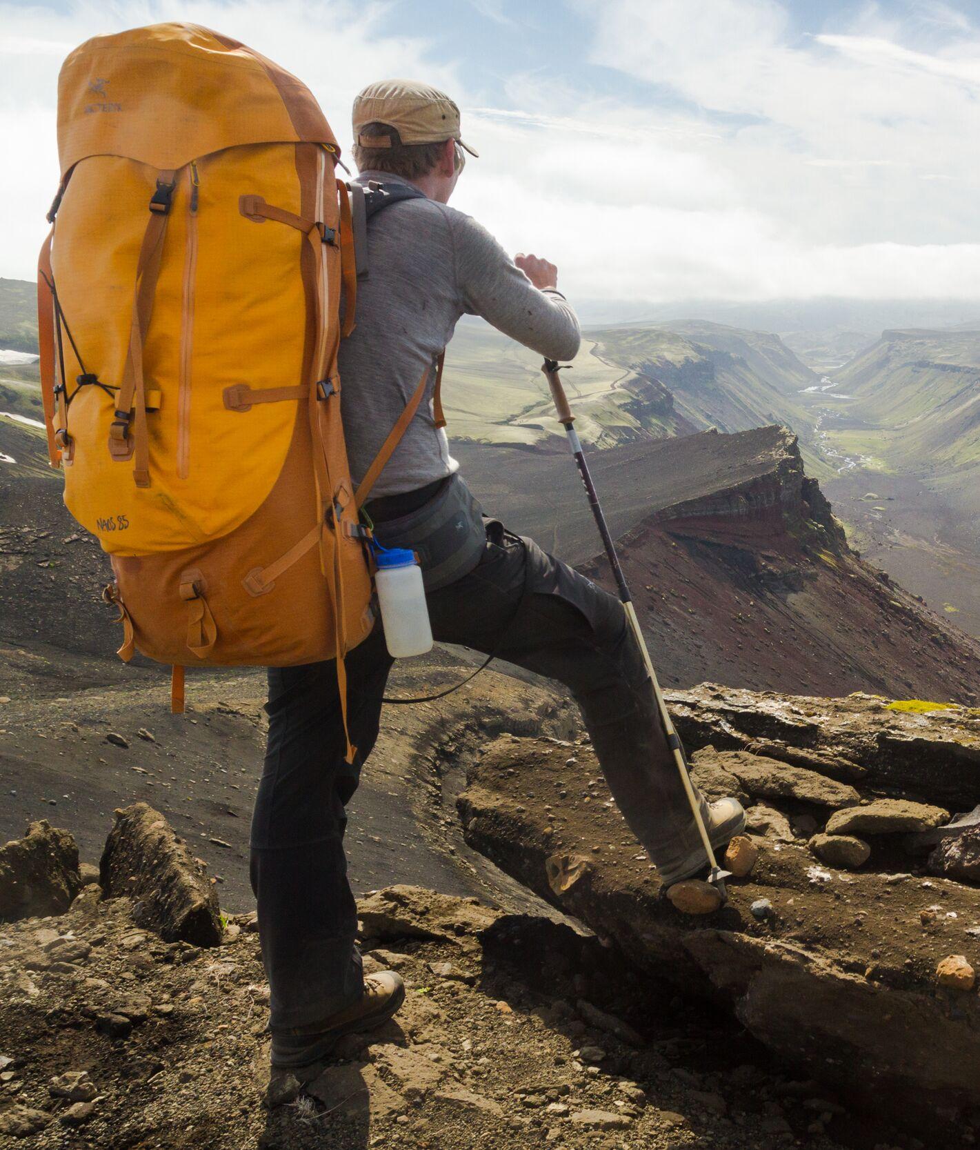 Hiker with a large orange backpack and trekking pole, standing on rocky terrain, overlooking a vast canyon in the Icelandic Highlands.