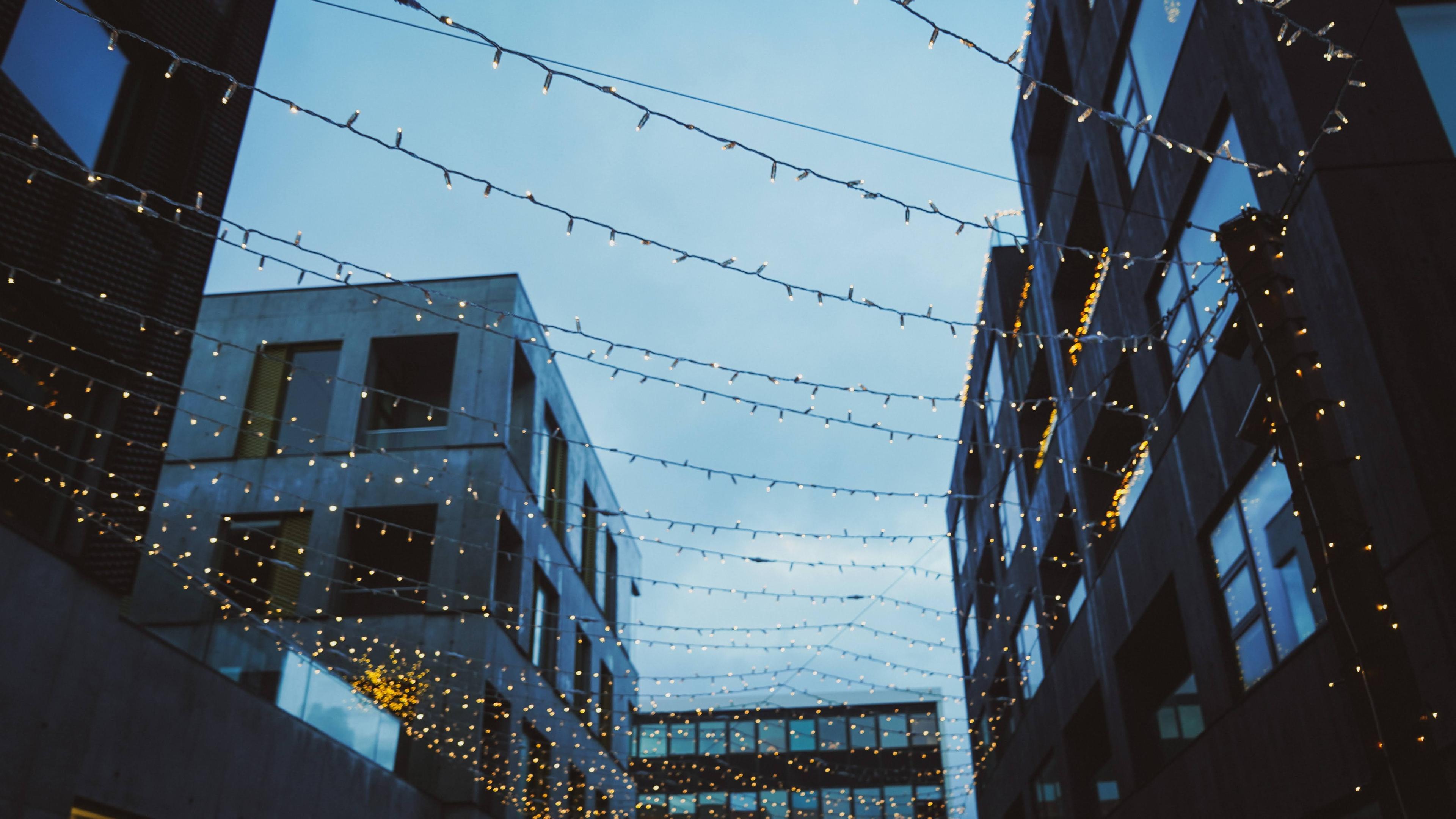 Fairy lights strung between modern buildings, creating a festive atmosphere in an urban setting at dusk.