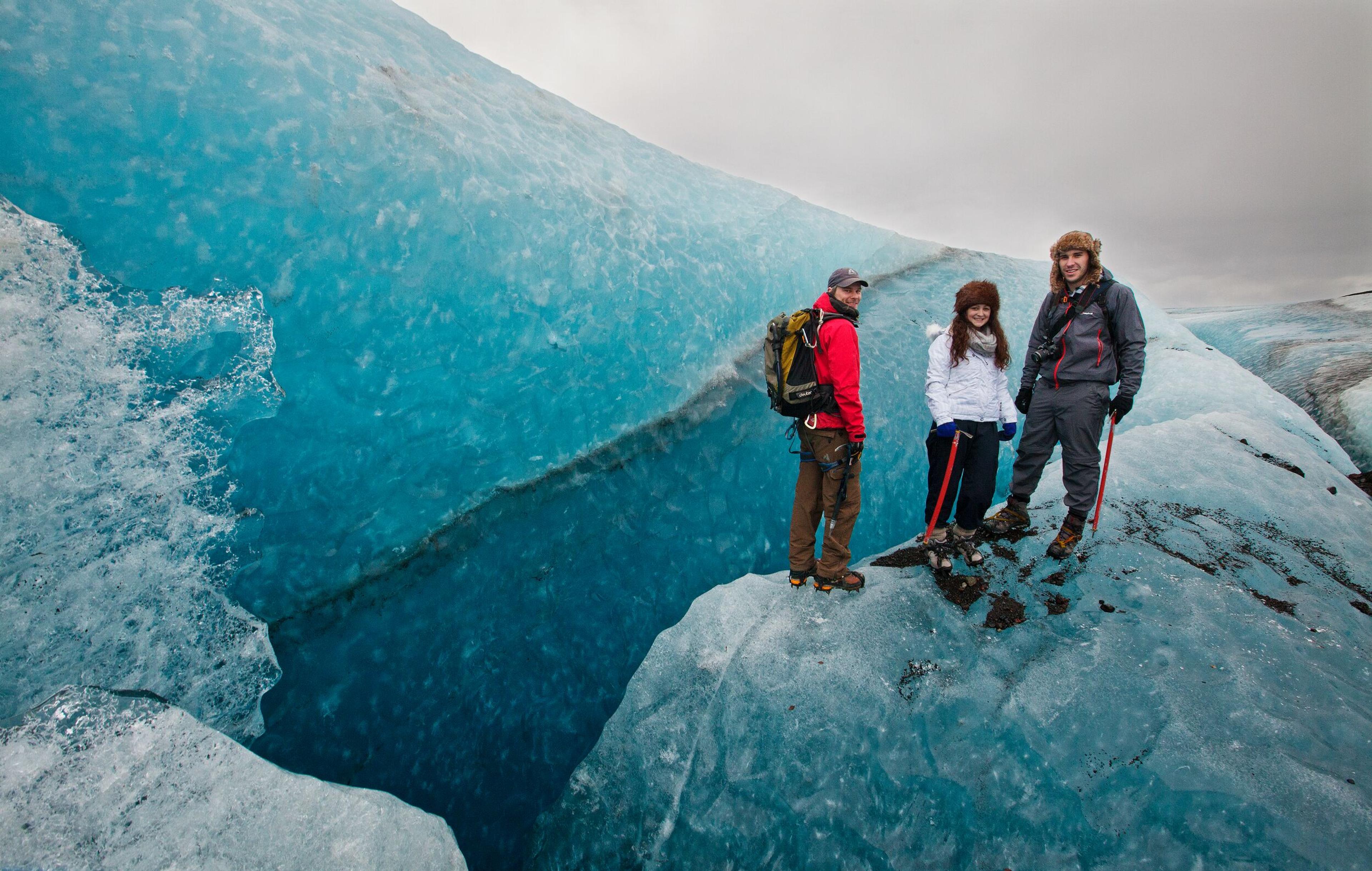 Three people standing on a blue glacier, next to a deep crevasse